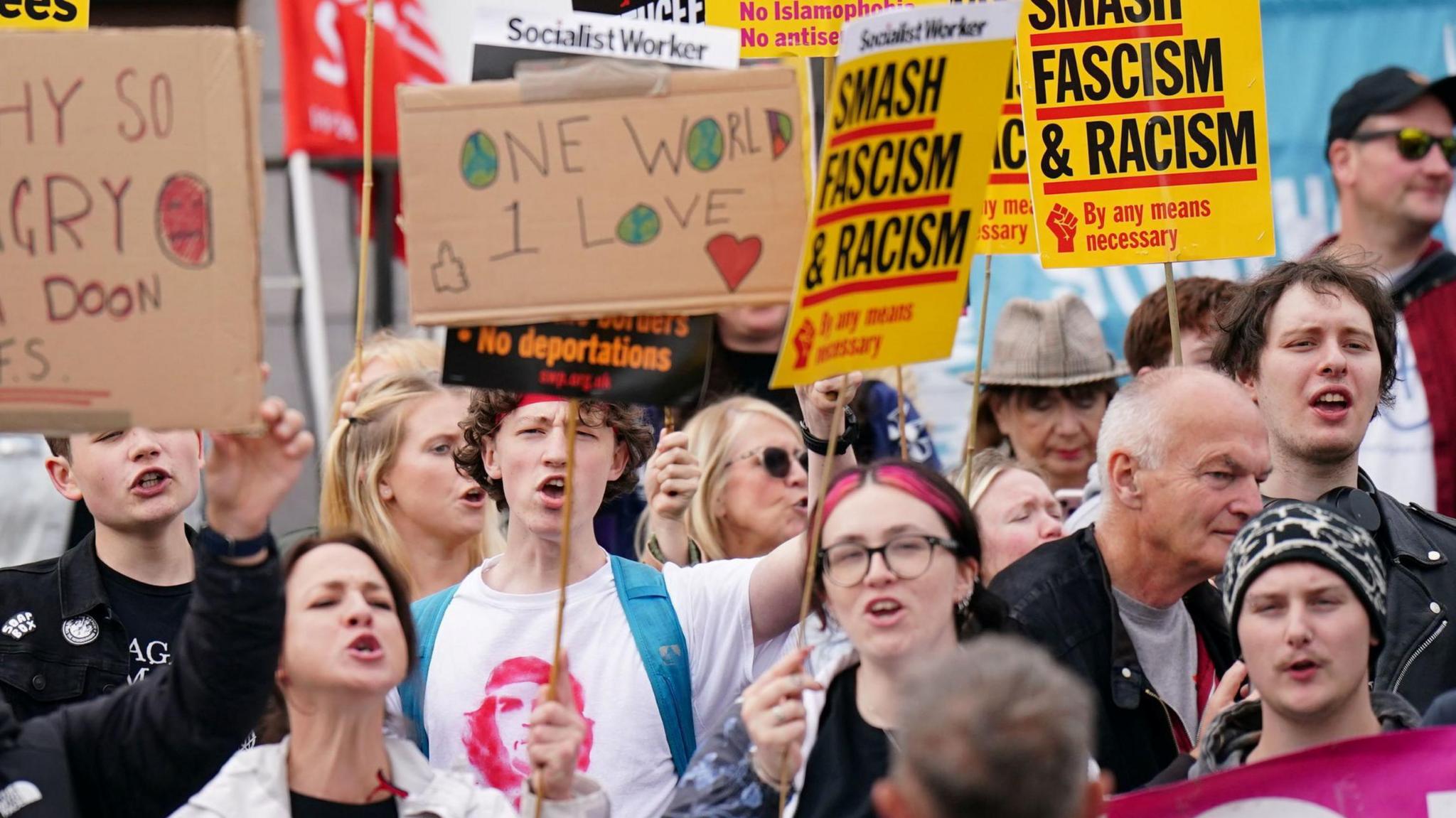 A group of pro immigration protesters stand together. It includes men and women. There are holding up another of signs. They read: "Smash fascism & racism" and "One world 1 love".