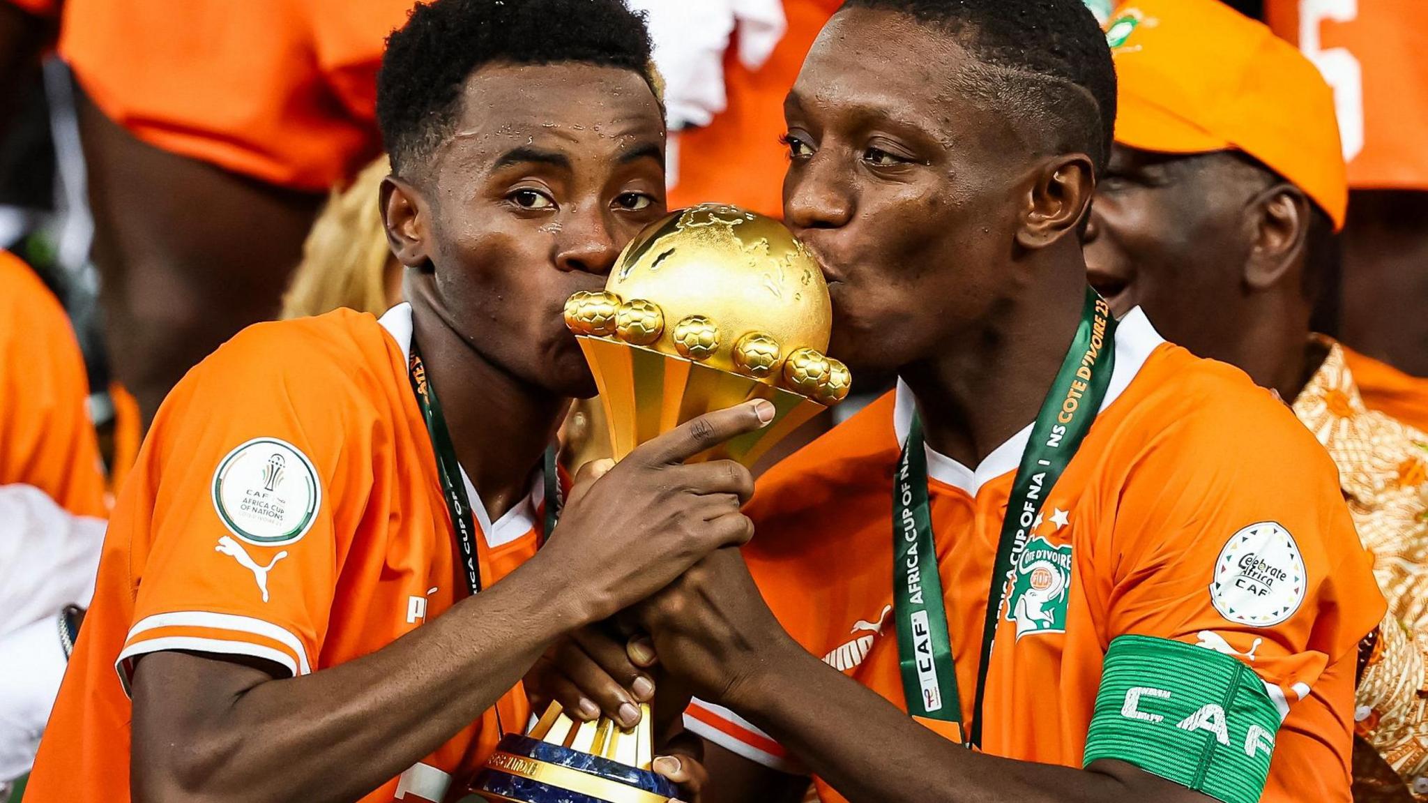 Simon Adingra and Max Gradel, wearing orange Ivory Coast shirts, kiss the Africa Cup of Nations trophy