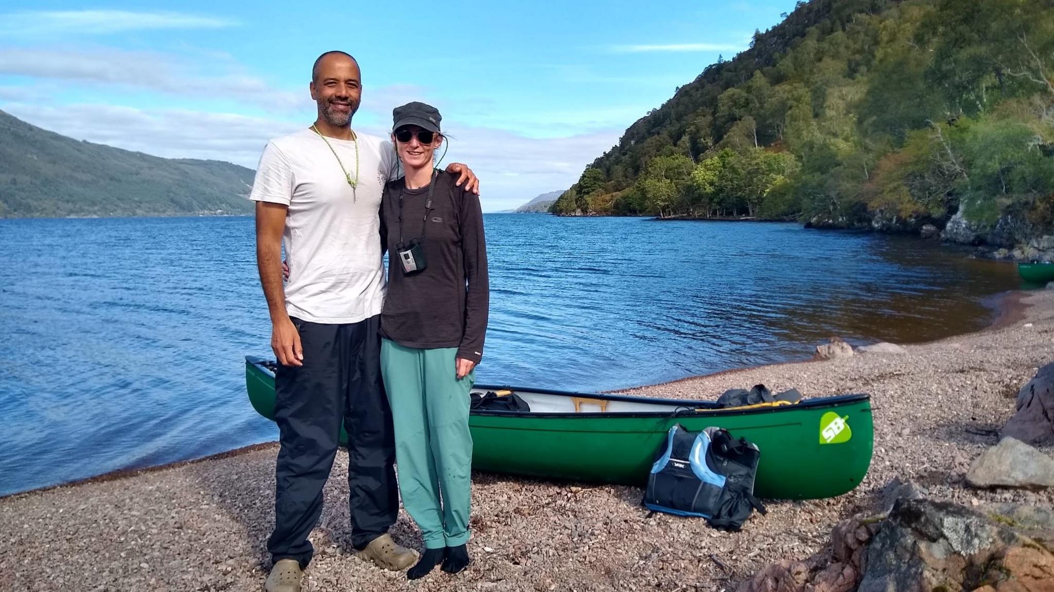 Tarig Sinada and Jenny Cawson. The couple are standing next to each other near a lake. A green canoe is on the shore