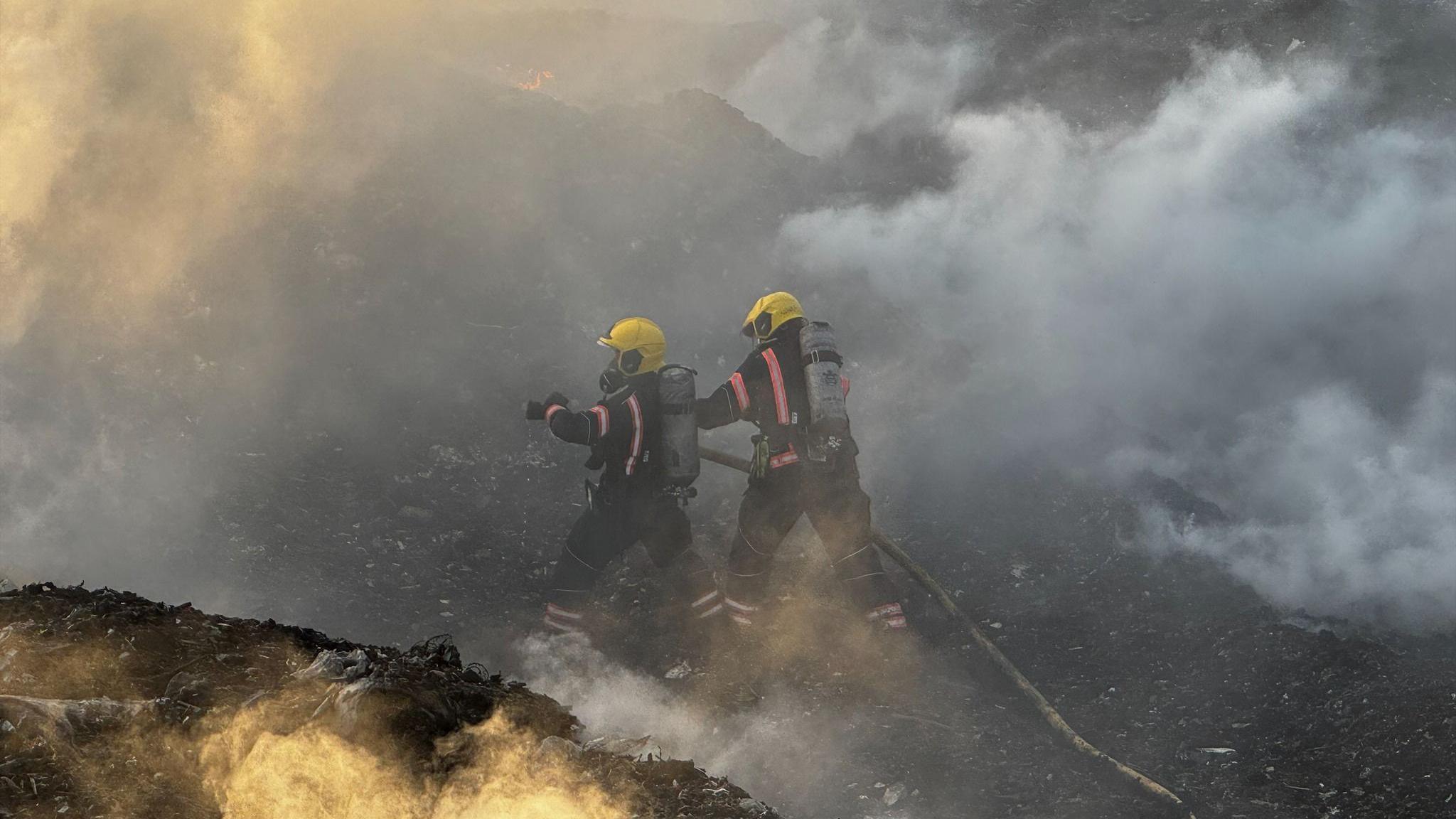 Two firefighters hold a large red hose to tackle a landfill site fire. They wear breathing equipment with tanks on their back as well as yellow helmets and black overalls.