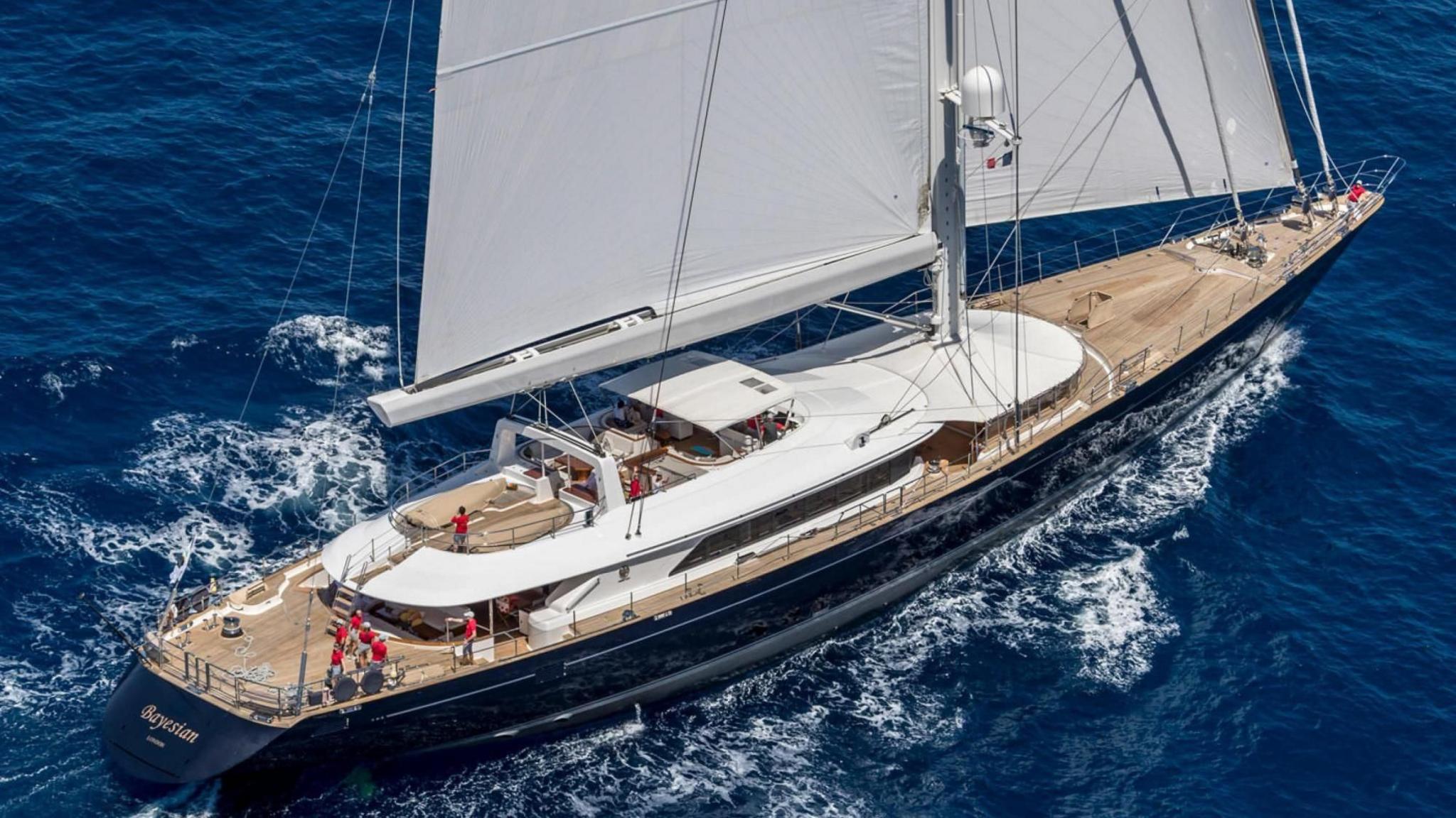 The Bayesian yacht with the lower parts of its white sails visible above the whole of its brown deck. The cabins have white roofs and the hull of the boat is black. About half a dozen people in red tops are visible on deck.
