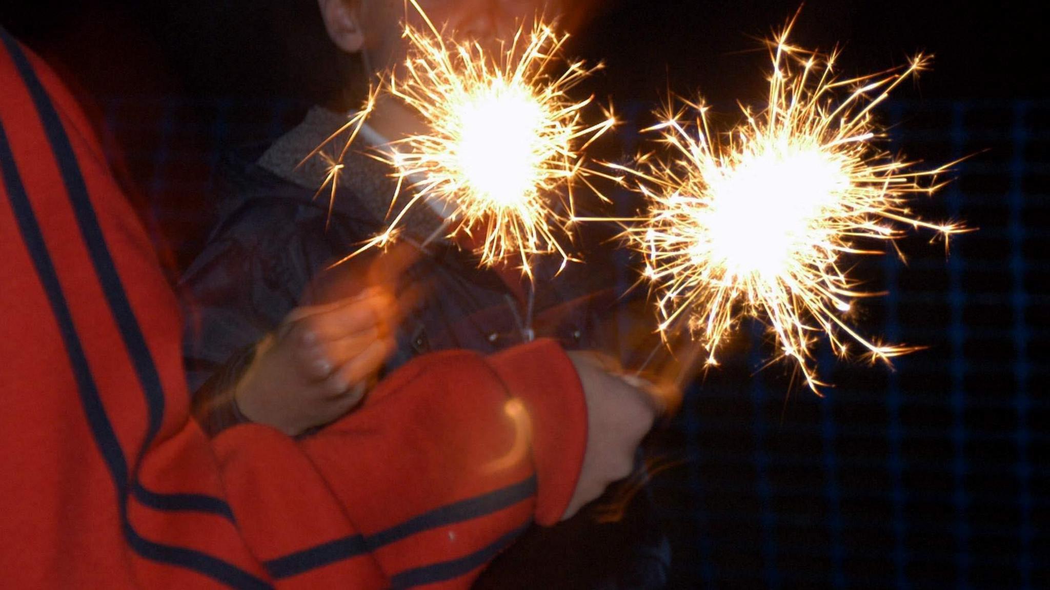 Two children are playing with sparklers; only their arms are visible and they are holding one sparkler each