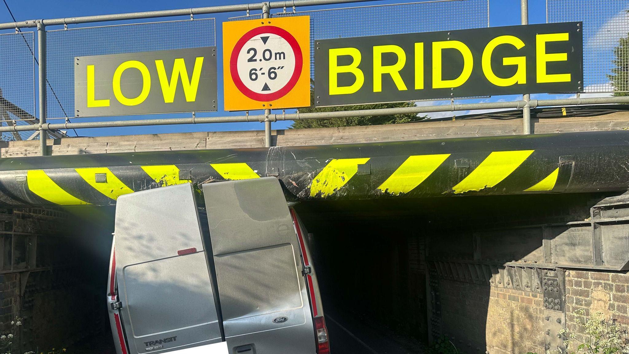 The back of a silver van which has become stuck under a bridge. The back doors have been wedged against the bridge - which has bright yellow stripes and yellow letters reading, Low bridge - 2.0m - 6'-6''.