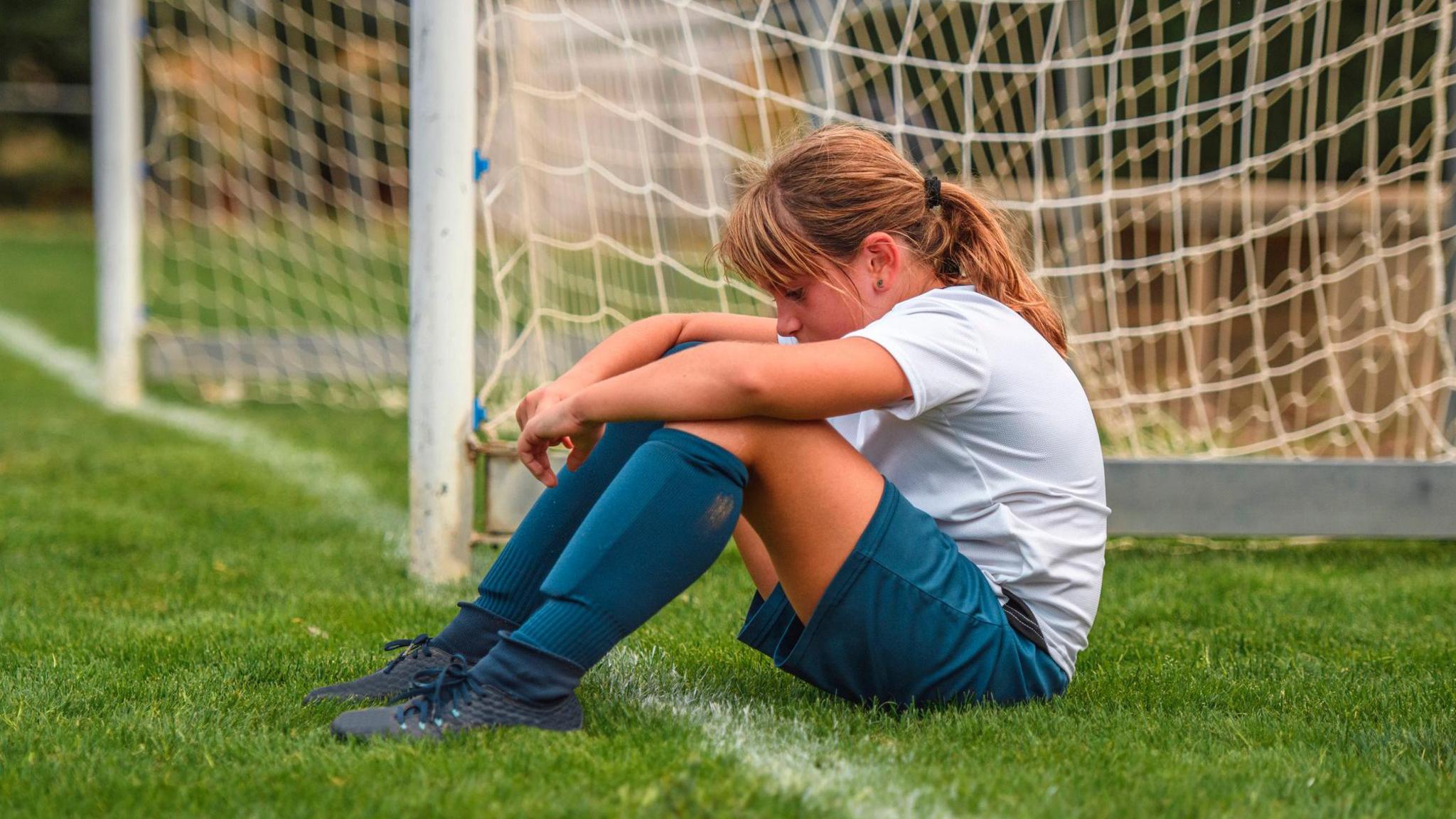 a girl sitting next to a football goal looking sad.