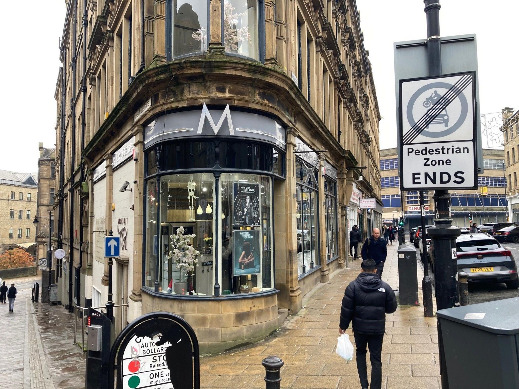 A circular shop window with black wooden frames on a street corner with Yorkshire stone pavements leading down both sides of the building. 