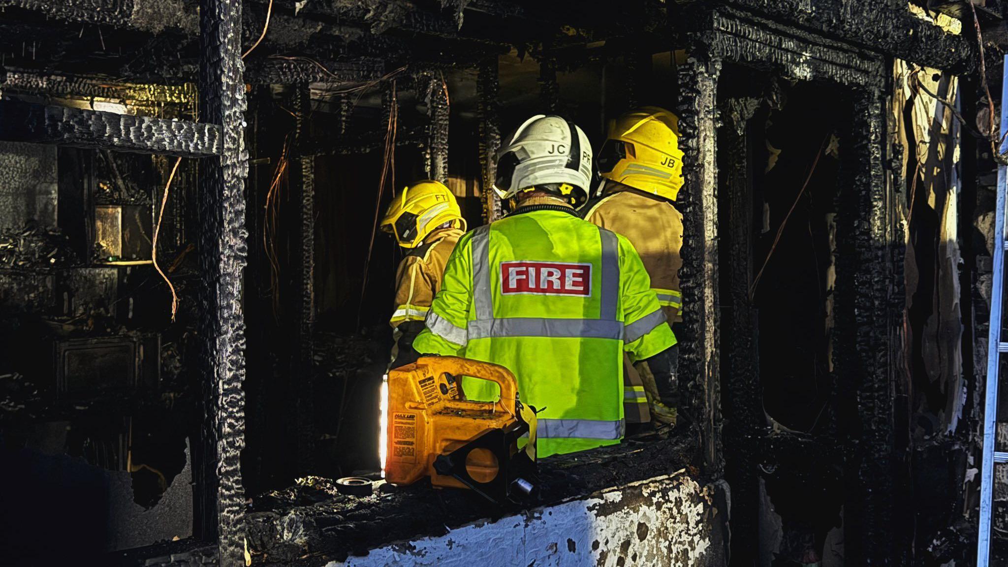Firefighters are standing inside a charred, burnt-out room. Everything is black after the fire and the firefighters are wearing hi-vis jackets.