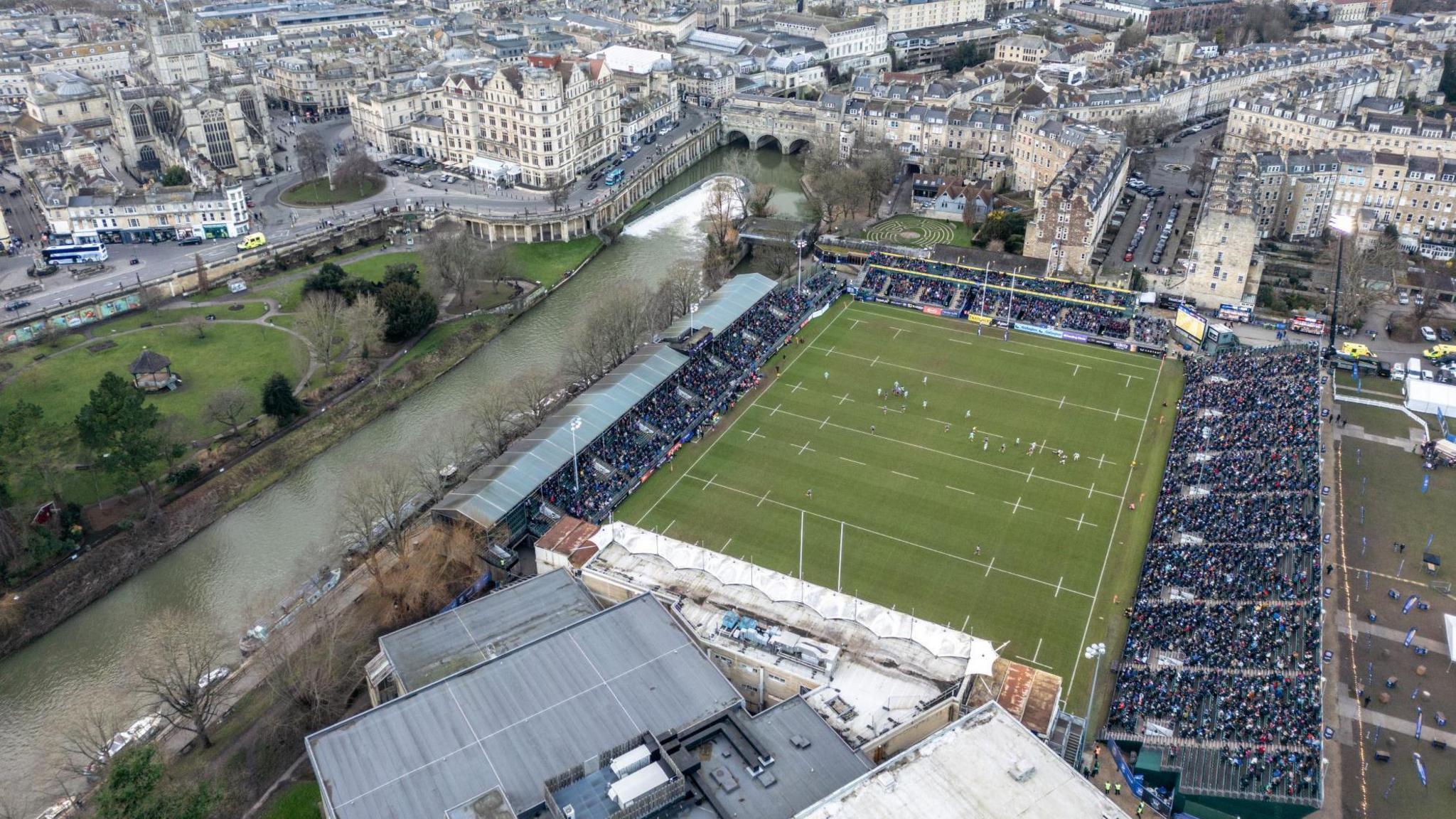 An aerial image shows the Recreation Ground, home of Bath Rugby, with the stands full. The image is taken on a clear day and much of the historic city centre is visible in the background