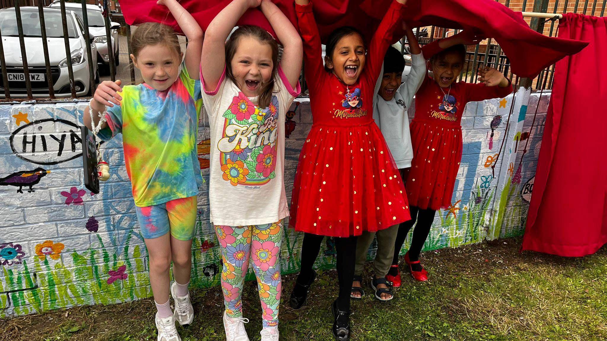 Five children named Jaylee, Mynah, Ashoka, Vishwan and Ananya stand in front of a mural they helped to paint. The mural is painted on a low wall that is topped with metal railings. The mural shows green grass at the bottom, a blue sky and buildings from Belfast's skyline. The children are holding up a red curtain that had been used to obscure the new mural before it was unveiled.