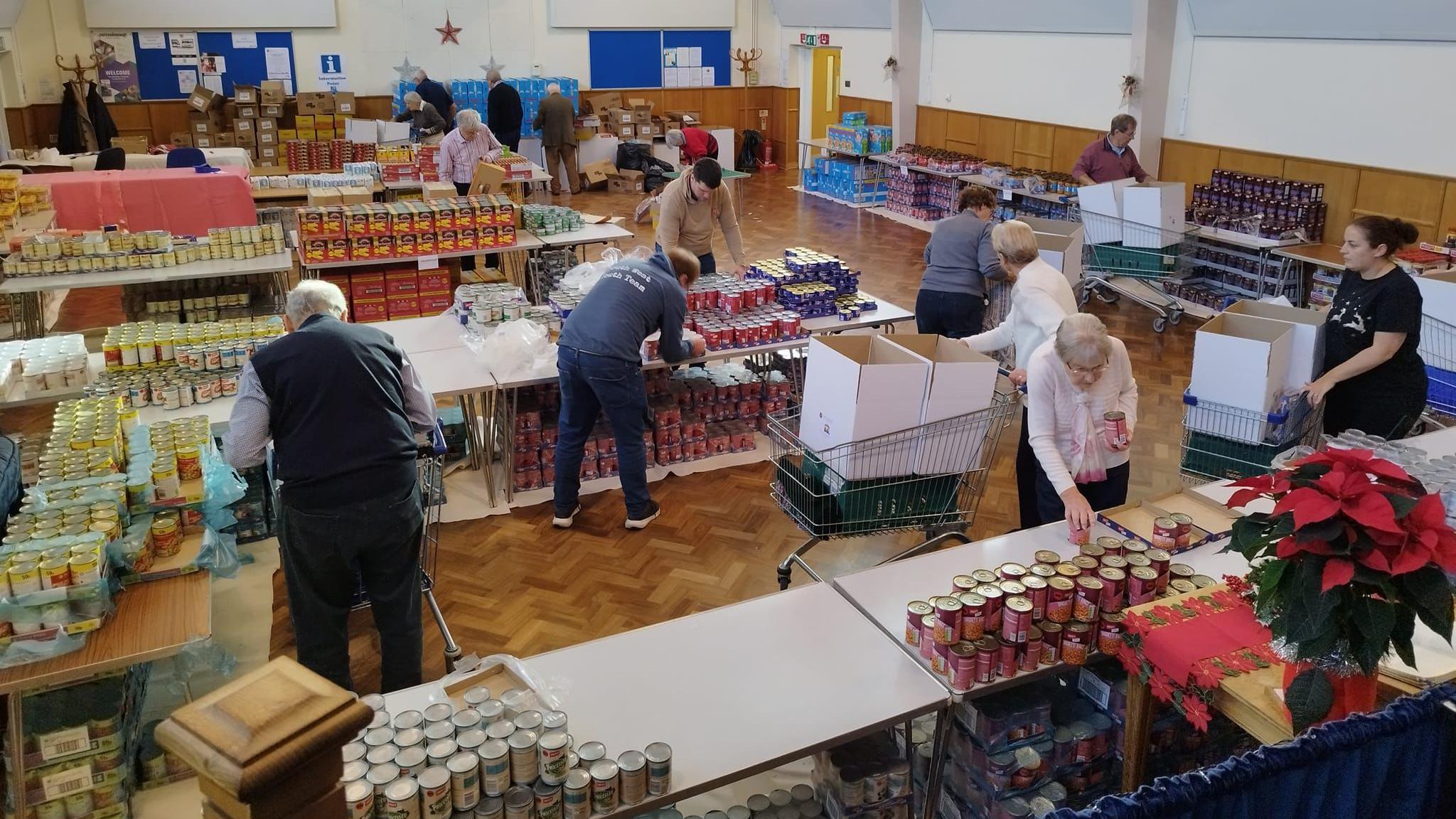 A group of 13 people stood in a large hall, filled with tables. The tables are stacked with tins, books and jars of food. The people are spread around the tables, packing boxes ready for the Salvation Army.