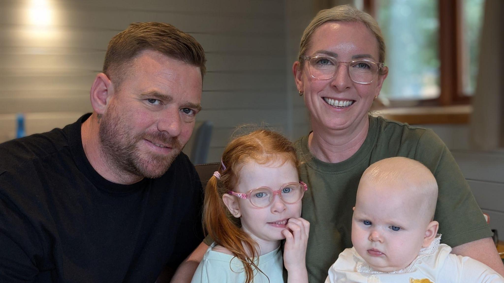 A middle aged couple holding young girl, who has ginger coloured hair and wears glasses, and a baby.