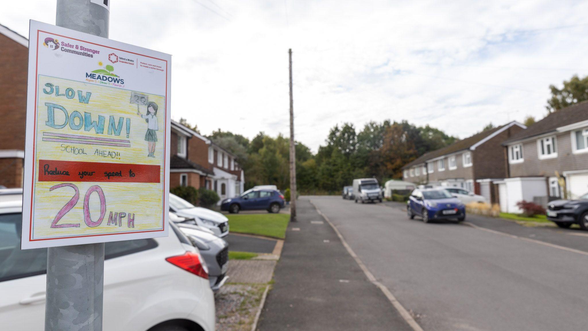 A road sign saying "slow down, school ahead" on a residential street, there are houses with cars parked in front of them on either side