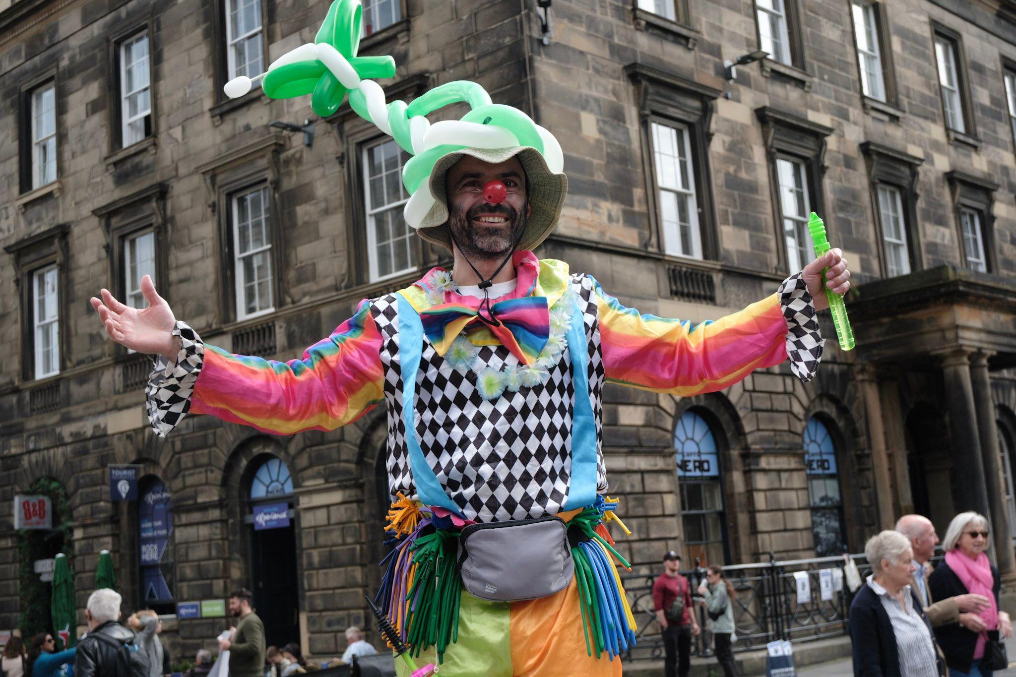A balloon model maker on stilts holding his hands wide as he smiles. He is wearing a green and white balloon hat, a checked and rainbow shirt, yellow and orange satin trousers and a red clown nose. He is standing on the corner of Princes Street with a big stone building behind him.