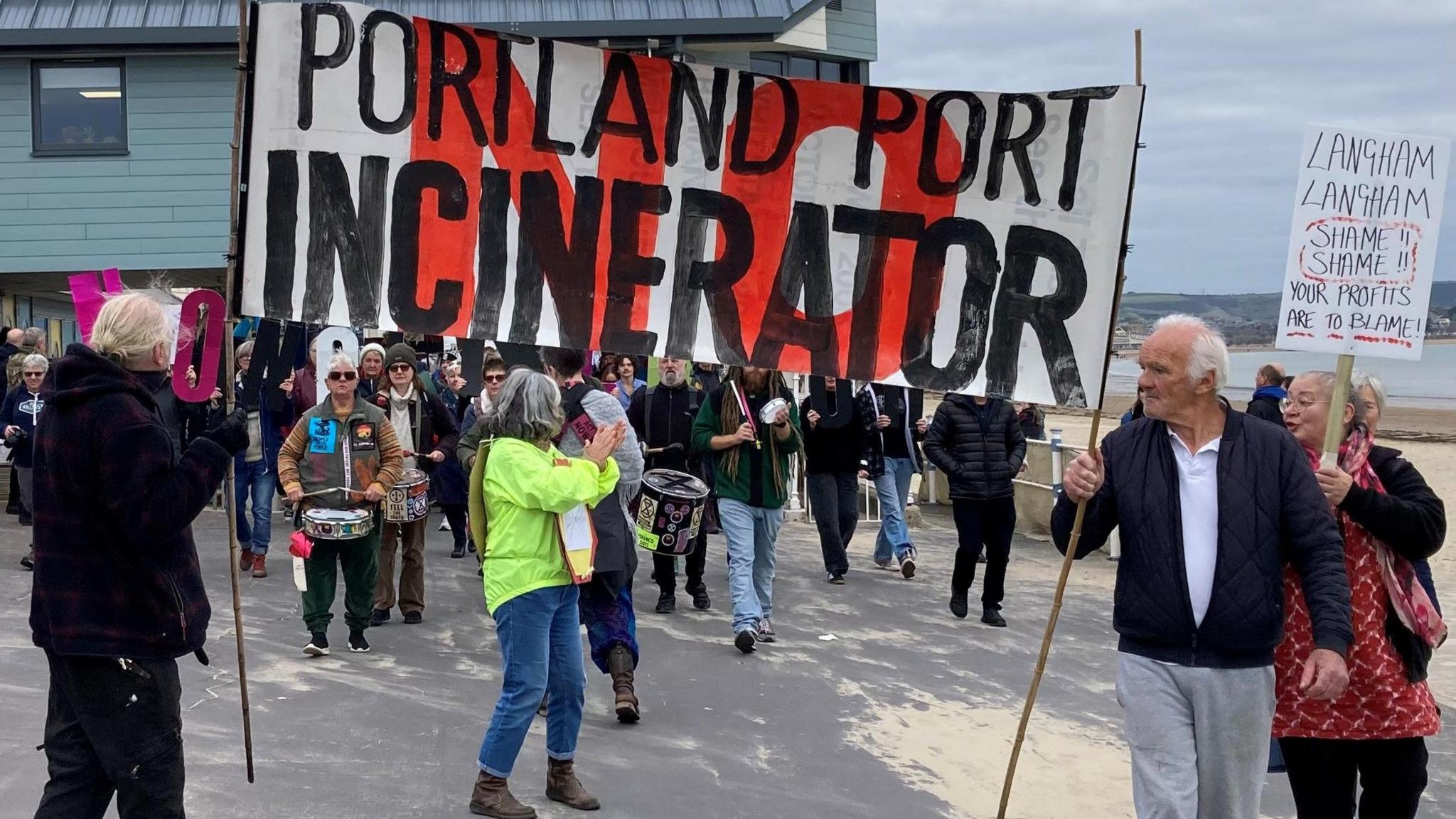 Two men with grey hair and wearing dark coats holding up a large sign saying 'no Portland Port incinerator". There's a crowd of people behind them, some holding up other signs and some playing drums.