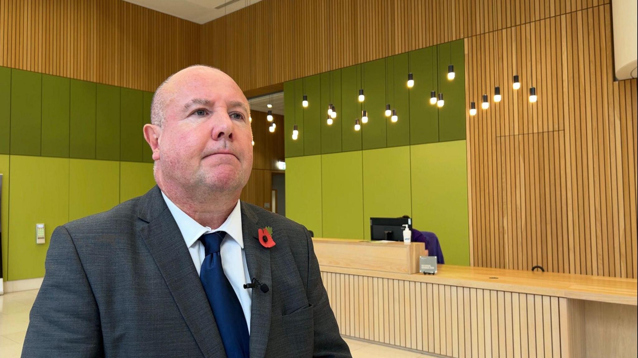 A man wearing a grey suit with a poppy in his buttonhole stands in a wood-panelled room.