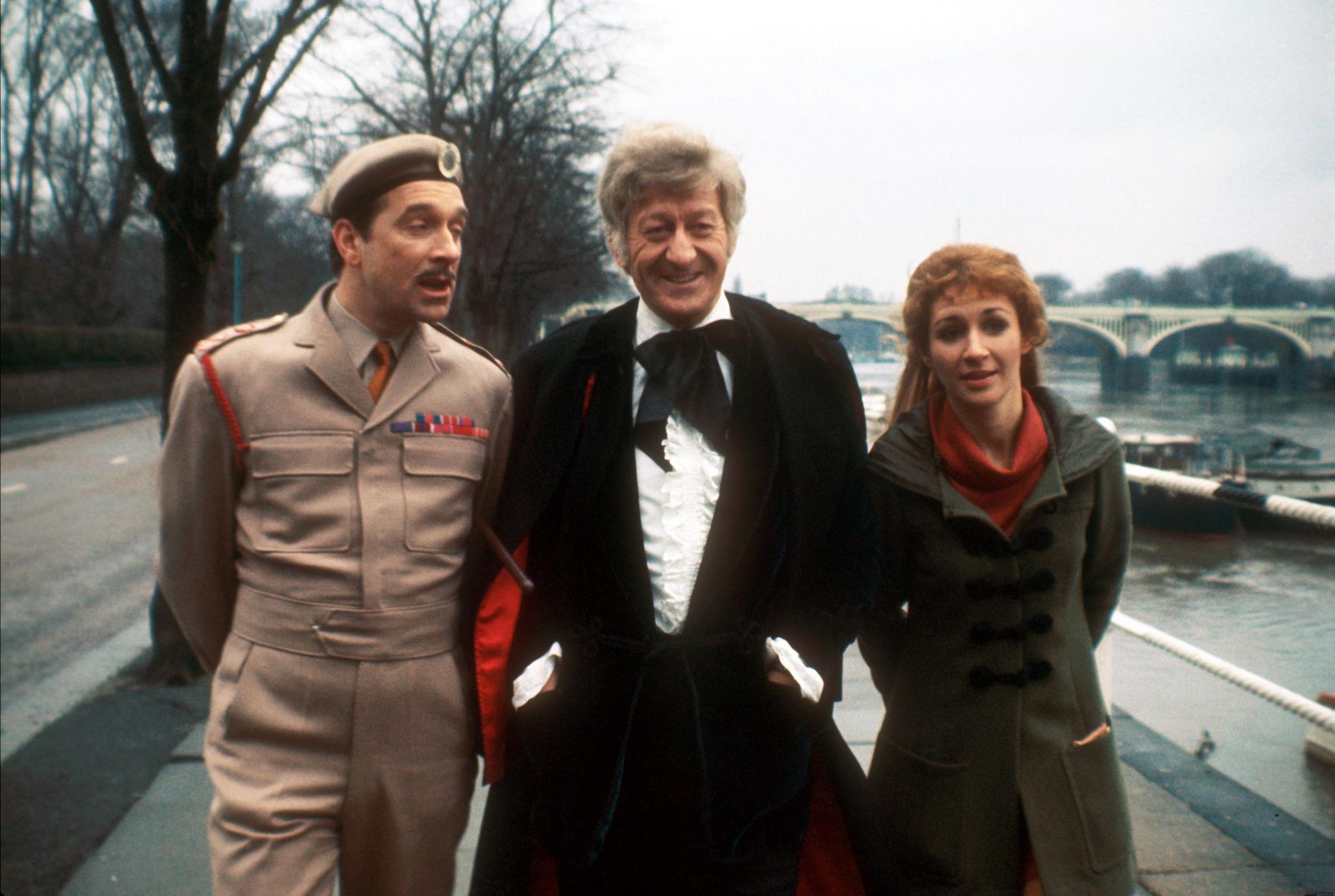 The 3rd Doctor is walking on a path alongside a river with Liz on his left and the Brigadier on his right. The Brigadier is wearing a beige military uniform with a red lanyard around his right shoulder and ribbons on the left side of his chest. He also wears a beret on his head with a UNIT badge above his left eye. Liz wears a dark grey coat with black toggle fastenings over a red dress. She has long blond hair pinned off her face, but with the length falling down her back. The Doctor is wearing a white frilly shirt with a black tie under a black jacket and cloak. The cloak has a bright red lining which can be glimpsed behind the Doctor, suggesting he’s moving.