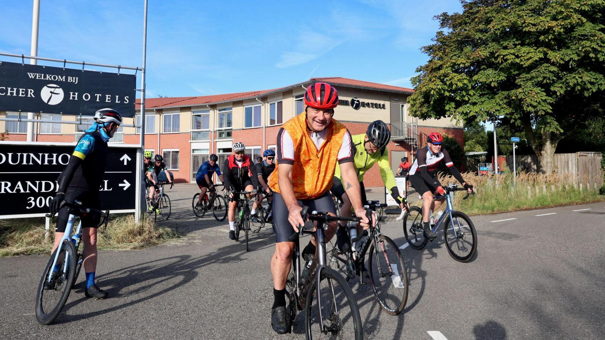 Men in brightly coloured Lycra on bicycles leaving a hotel. They are all wearing cycle helmets.