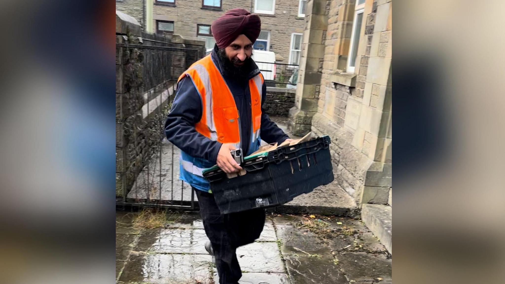 Bill Mato carries a box of aid into the stone-built Trallwn Community Centre in Pontypridd. Terraced houses can be seen behind him and there is water on the pavement.