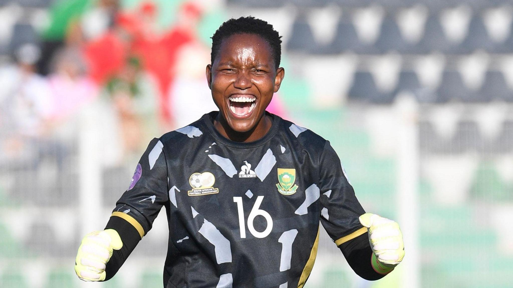 Andile Dlamini wears a black South African goalkeeping jersey with grey trim along with white gloves, as she clenches her fists and opens her mouth wide to scream towards the camera