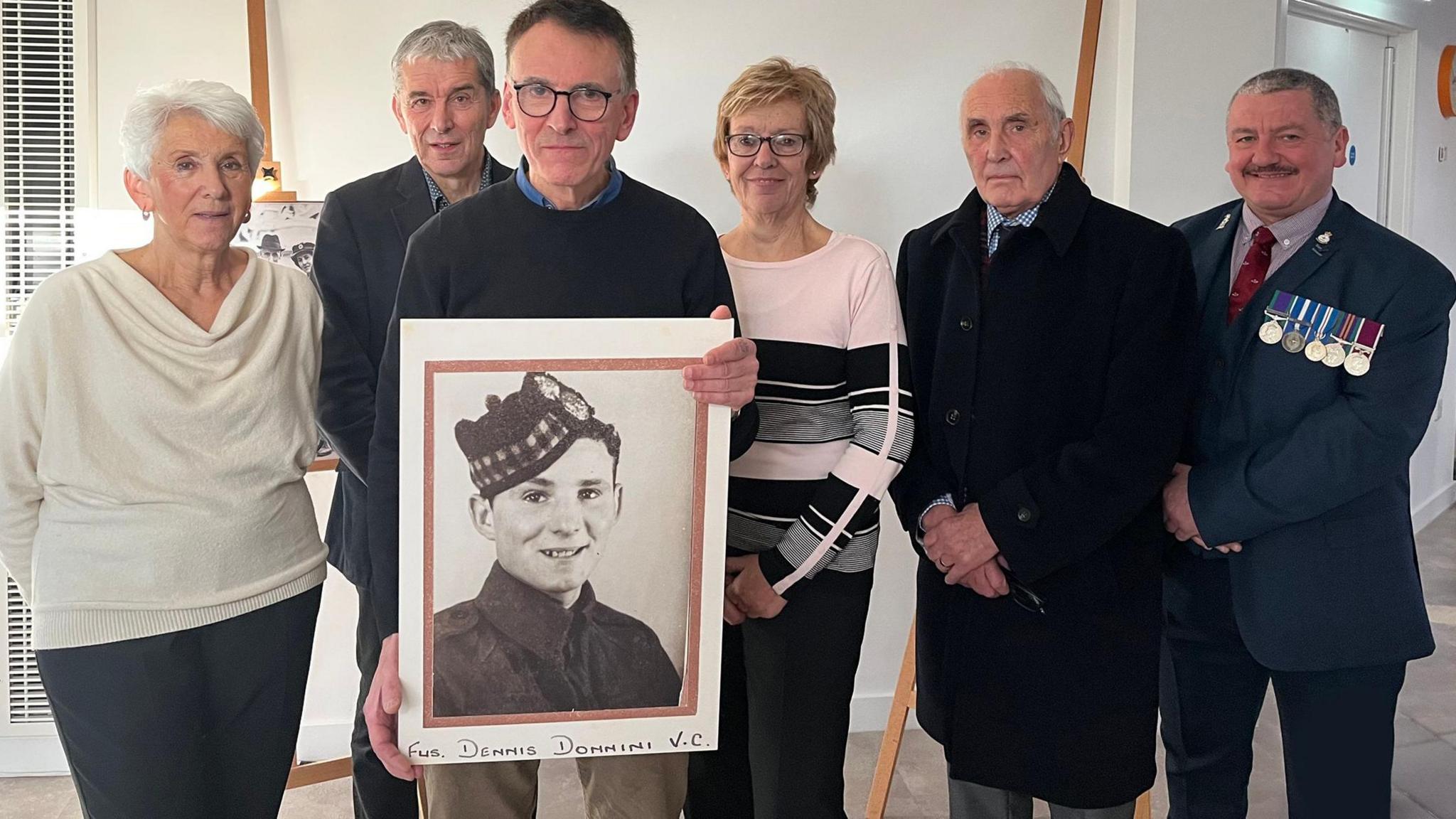 Group of six people posing for photo. Jerome Donnini is second from the right. There is also a man wearing a suit and medals on his breast-pocket. 
Two other women and three men, are dressed smartly. The man in the middle is holding a large photo of Dennis Donnini wearing a military hat. 