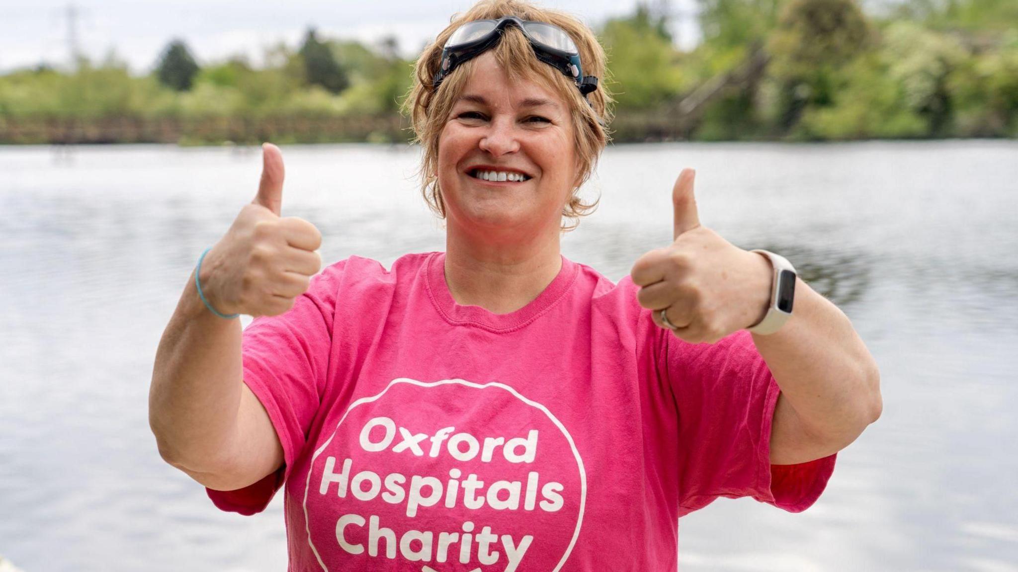 A smiling Sarah Vaccari by the pool, giving two thumbs up for the camera. She is wearing a pink T-shirt with the Oxford Hospitals Charity branding and goggles on her head.