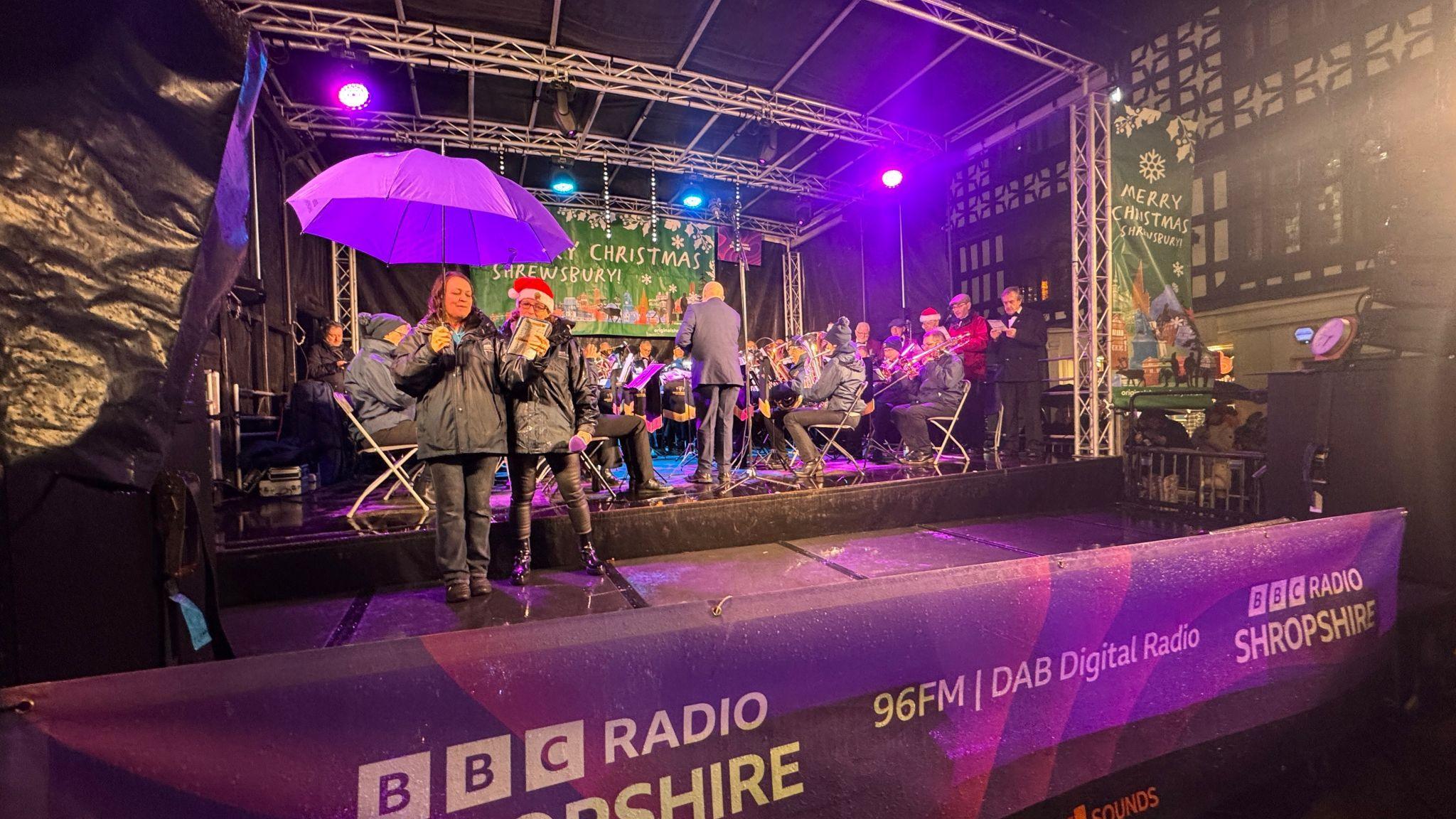 Two people under a purple umbrella and wearing coats, stand in front of a group of seated musicians playing a range of instruments. They are on a stage and at the back is a poster with "Christmas" and "Shrewsbury" on it.