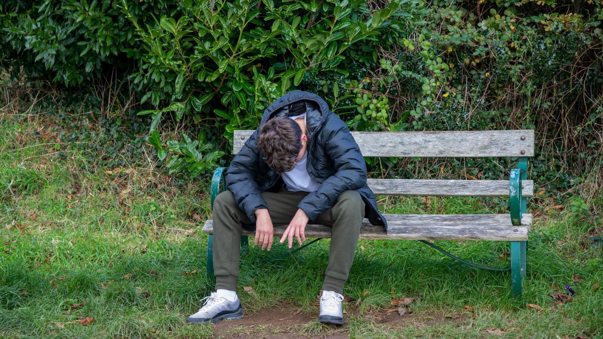 A youth in a coat and tracksuit bottoms sits on a bench in a park with his head bowed towards the ground