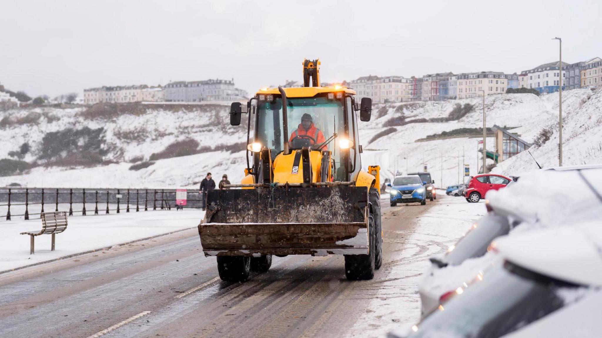 Digger drives on road toward camera. The landscape surrounding it is very snowy, with cars parked to the right of the picture topped with snow. The road is covered in a brown slush.