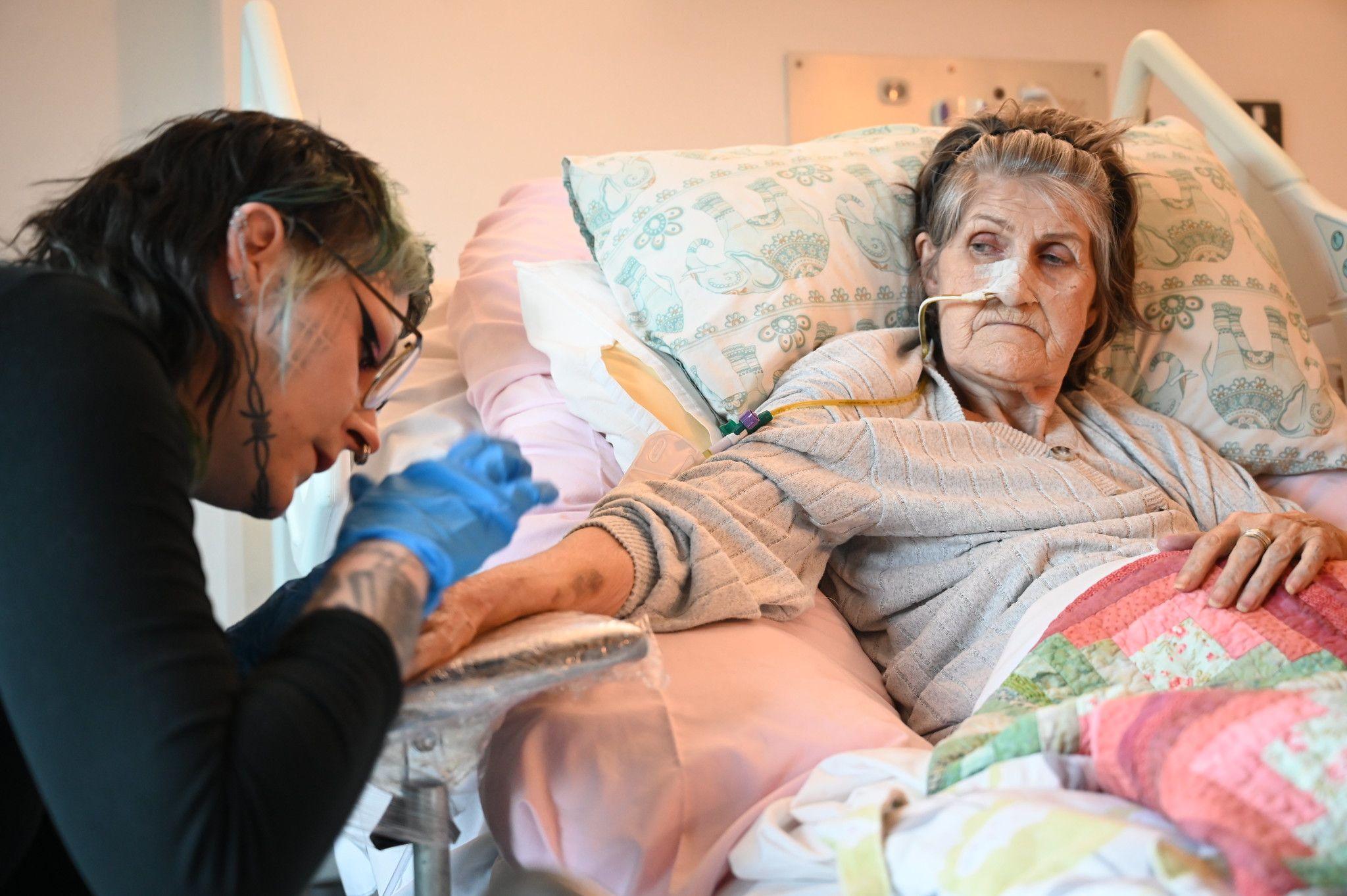 An elderly woman lies in a hospital bed, with a tube coming out of her nose. A younger woman wearing blue gloves is tattooing her forearm.