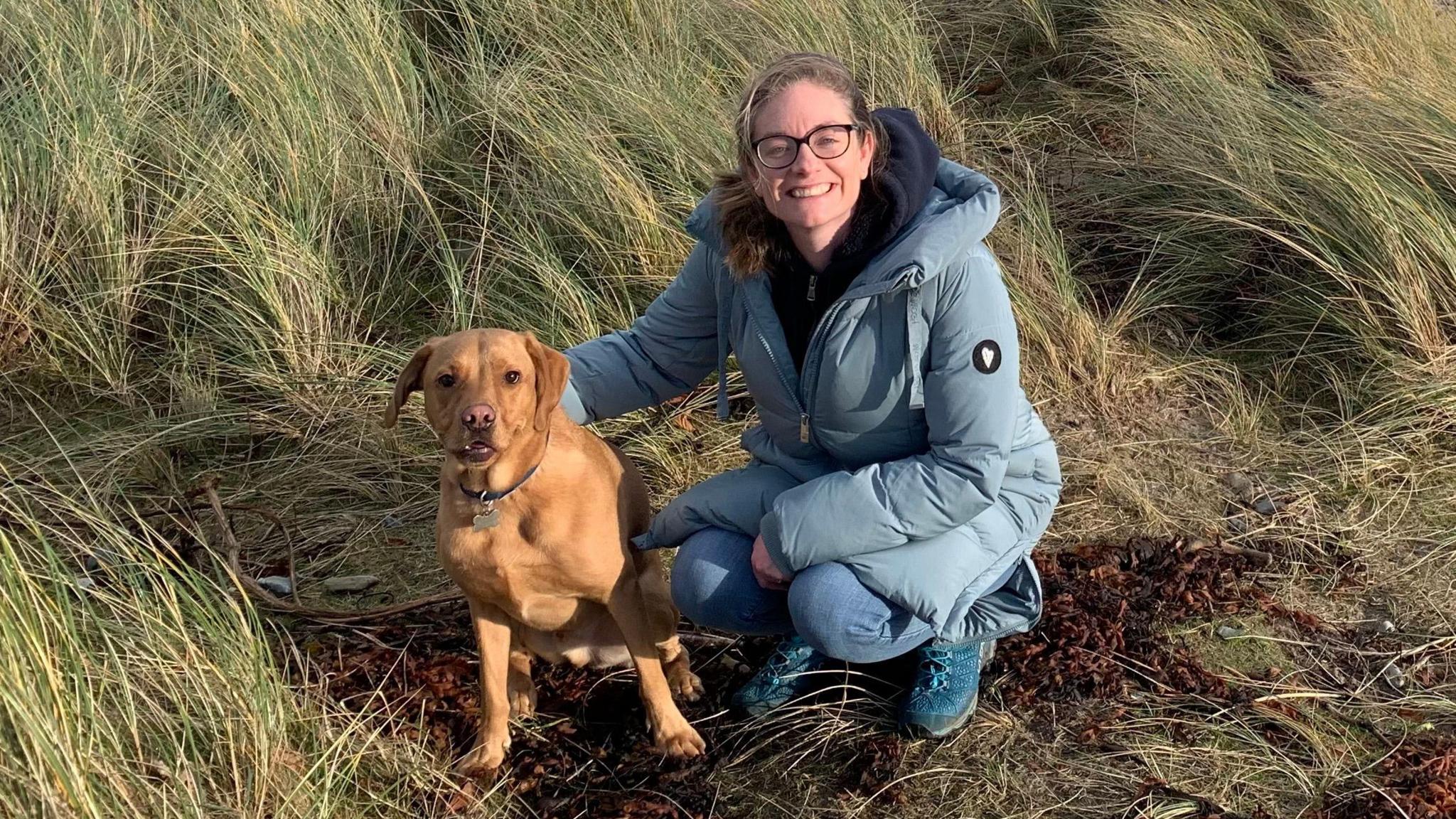 A woman wearing a long light blue coat and blue trainers crouches beside her golden labrador dog.  They are looking into the camera and are on a grassy area beside the sea.

