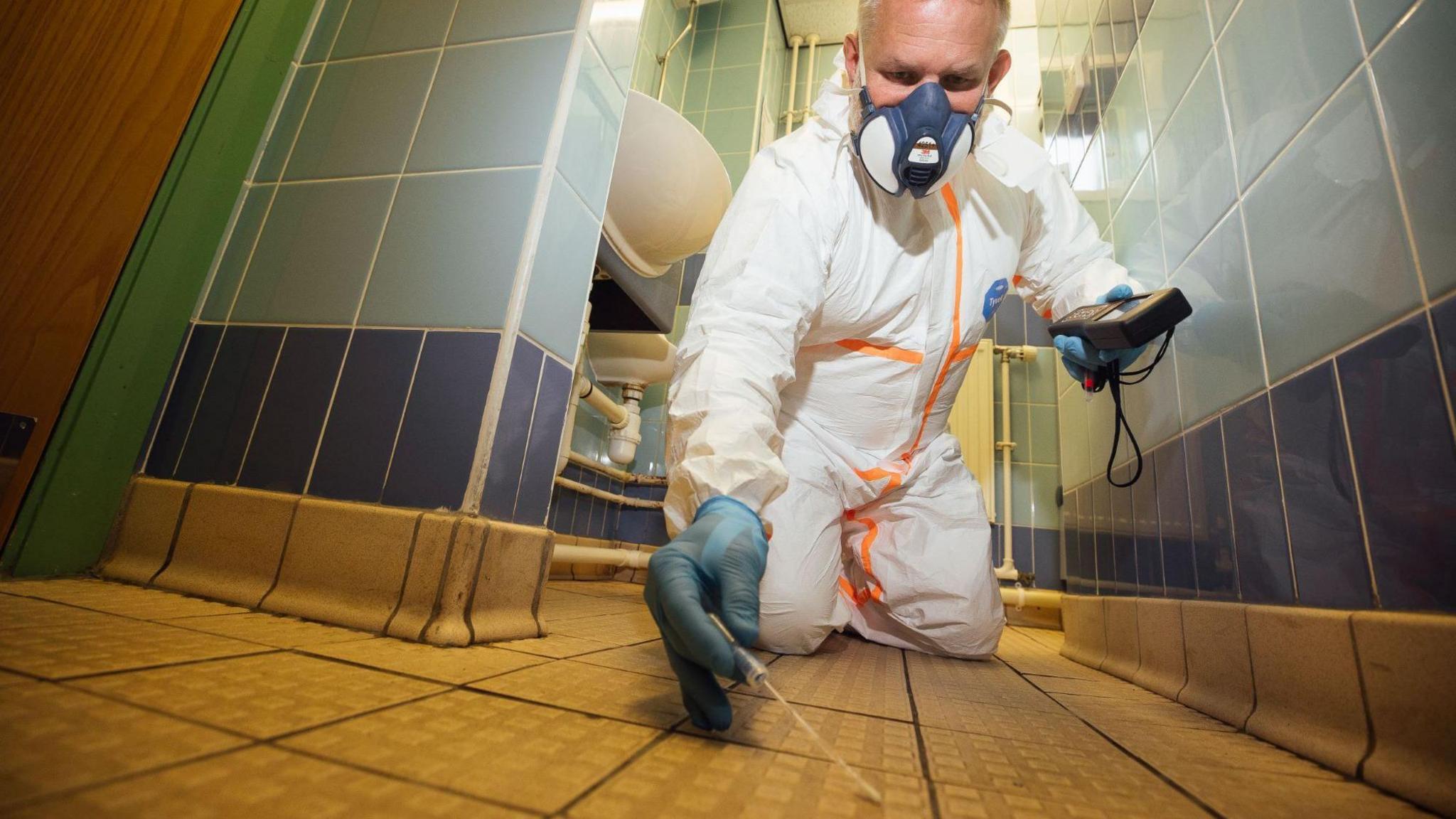 Ben Giles photographed kneeling over inside a blue tiled bathroom. He wears full white PPE, blue latex gloves and holds a cotton swab against the bathroom floor tiles. In his other hand he holds a black device. He wears a face mask and has grey short hair. In the background there are two white sinks.