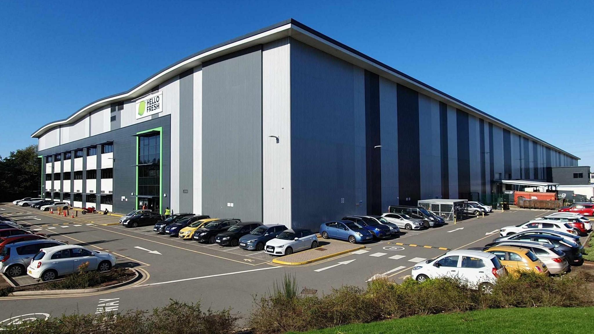 A large grey and white warehouse with the Hello Fresh logo above the main entrance, surrounded by a several rows of cars in the company's car park.