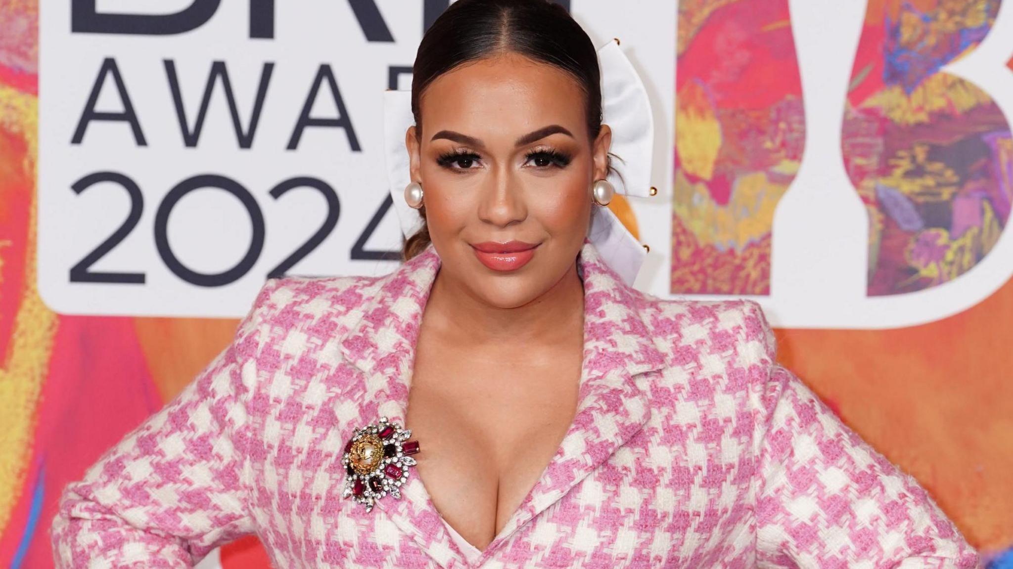 Head and shoulders shot of Rebecca Ferguson in a pink dogtooth jacket standing in front of a Brit Awards banner