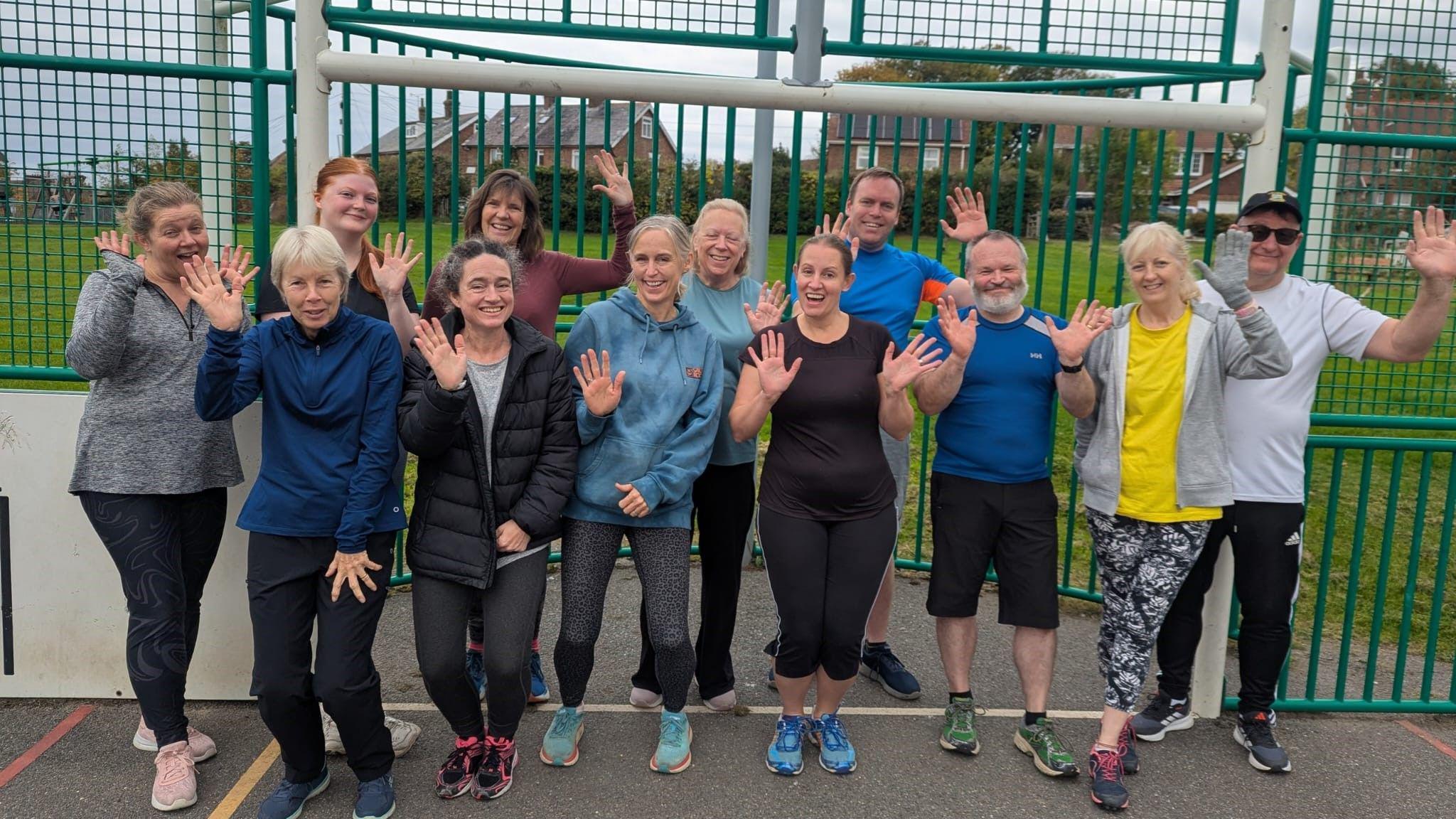Twelve people, a group of men and women, all wearing workout clothing, pose on a hard exercise surface in a park, waving to the camera.
