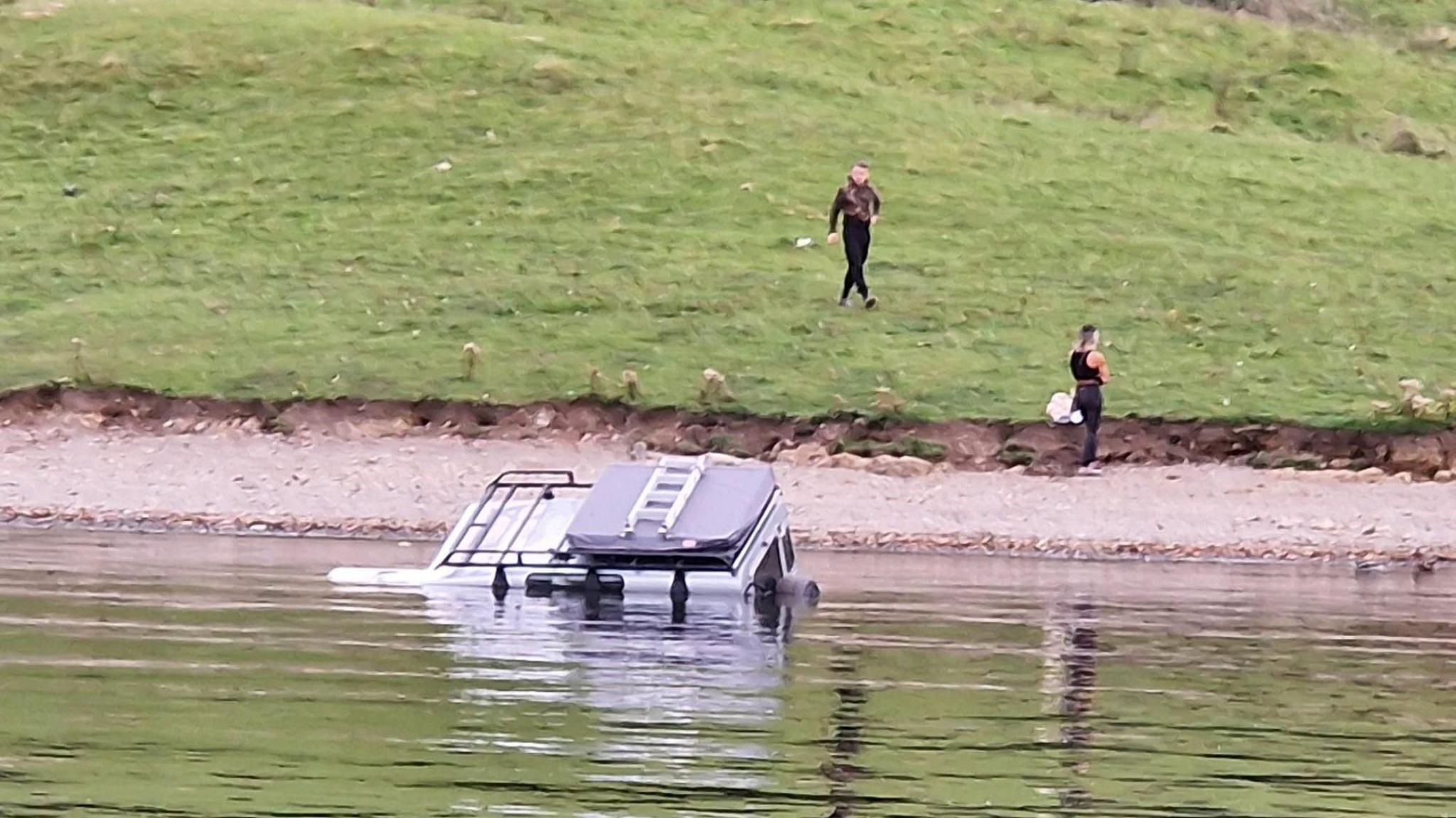 The Land Rover almost totally submerged in Windermere with two bystanders on the nearby shore.