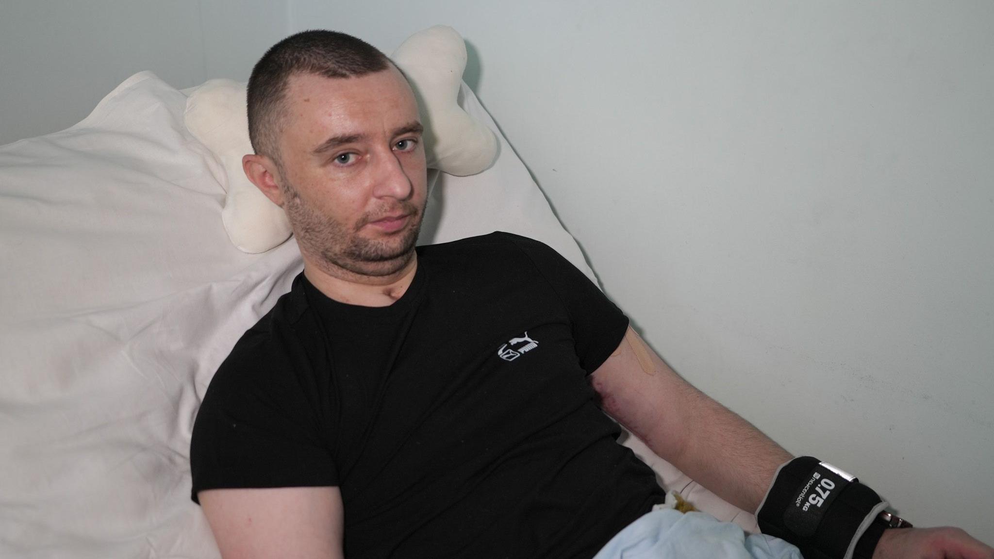 A man wearing a black T-shirt and with a scar on his neck from a tracheostomy sits in a hospital bed