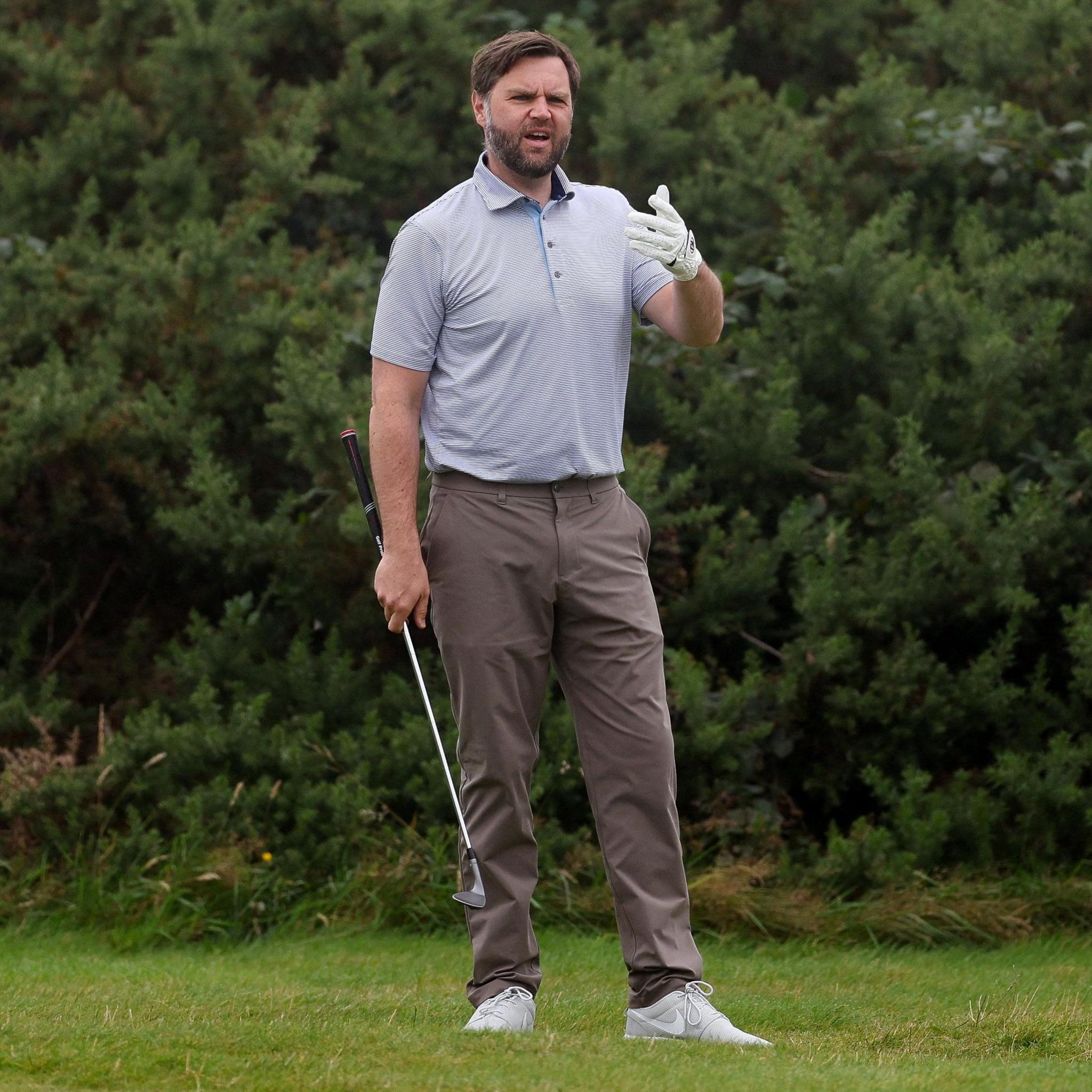 A man with brown hair and bear, wearing a light blue top an brown trousers holds up a gloved hand while holding a golf club