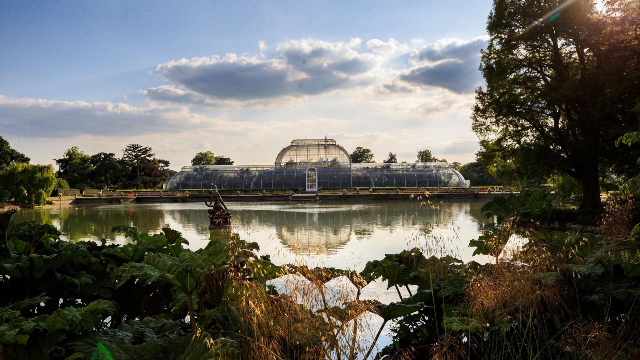 A view across a lake towards a large green house with a central two story dome and single-story wings to either side. The green house is reflected in the lake. Trees and shrubs stand in the foreground. The sky is blue.