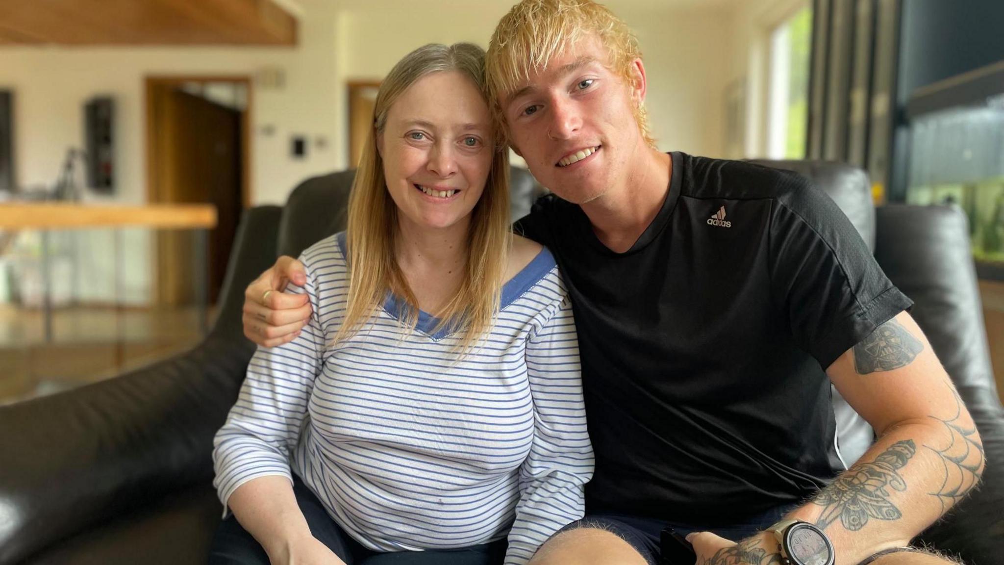 A young blonde haired man with tattoos sits with his arm around an older woman with a striped t shirt.