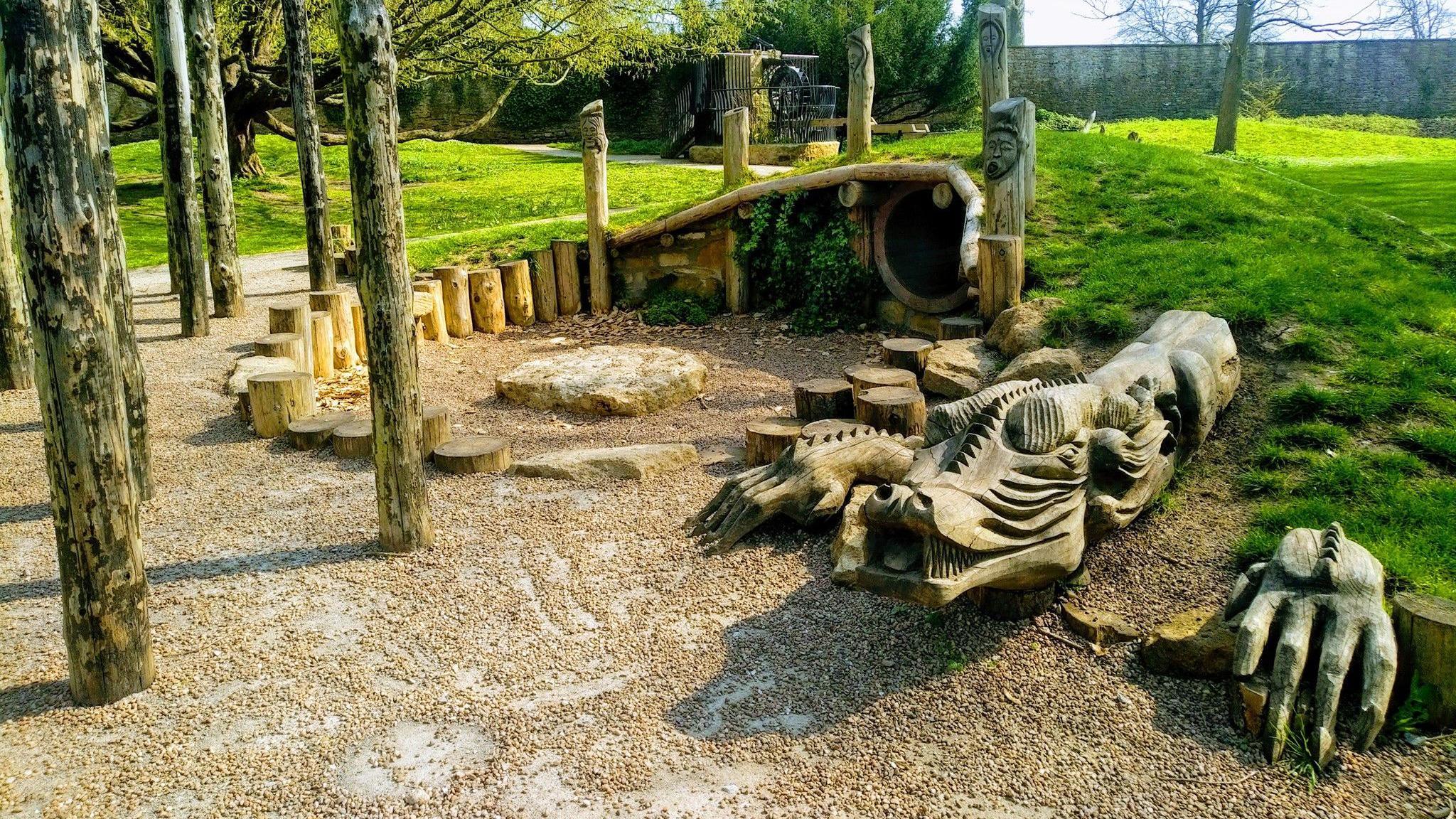 A feature at a play park that is shaped like a dragon. It is made of wood. There is green grass around it.