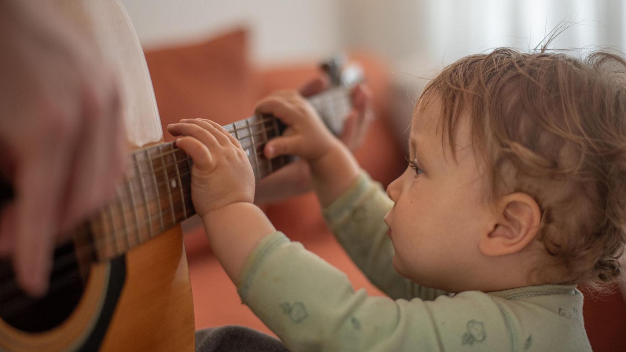 A toddler reaches up to strum the strings of a guitar, being held by an adult
