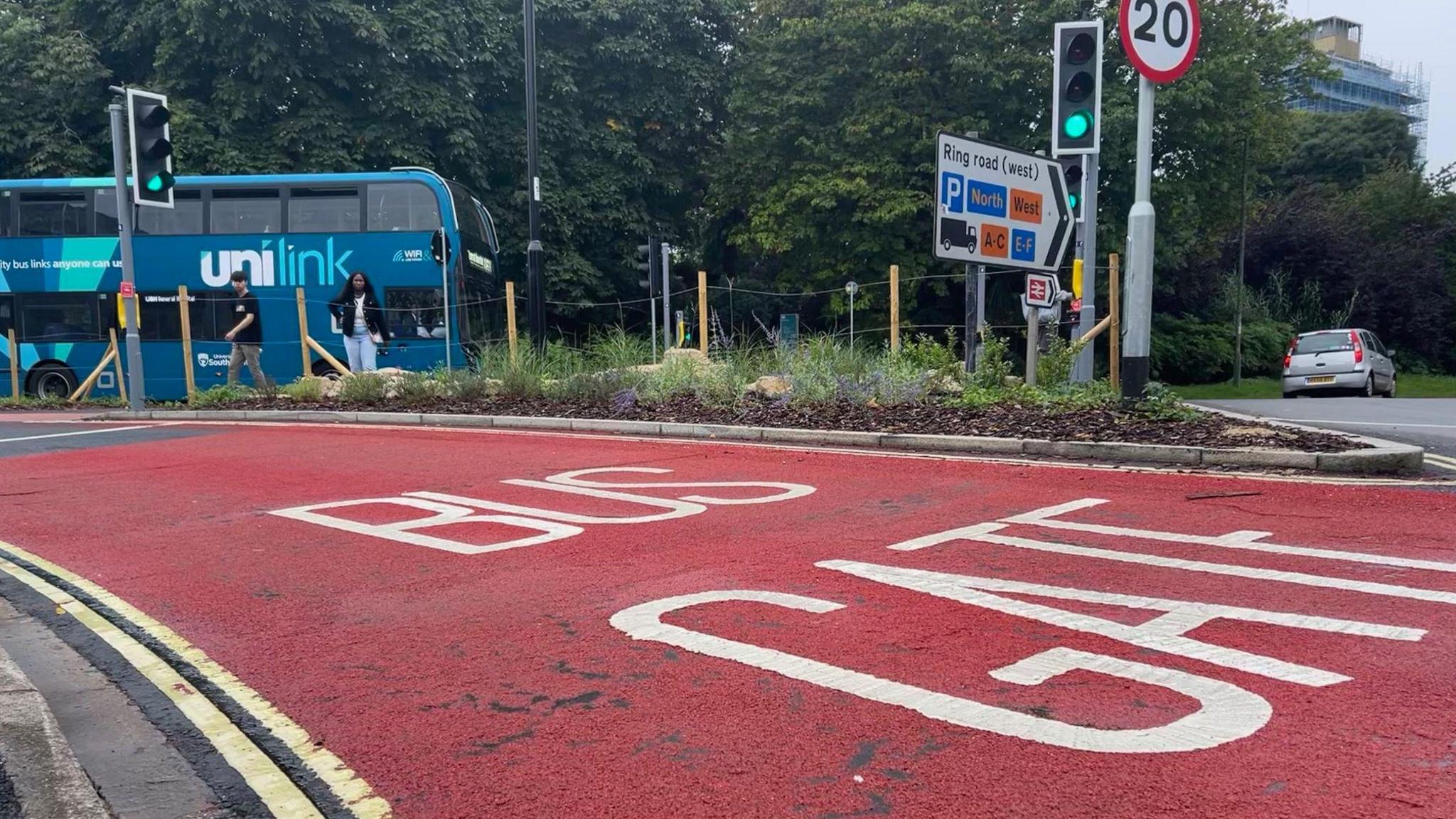 A road painted red with the words 'BUS GATE' in large white writing. In the background there is a double decker bus.