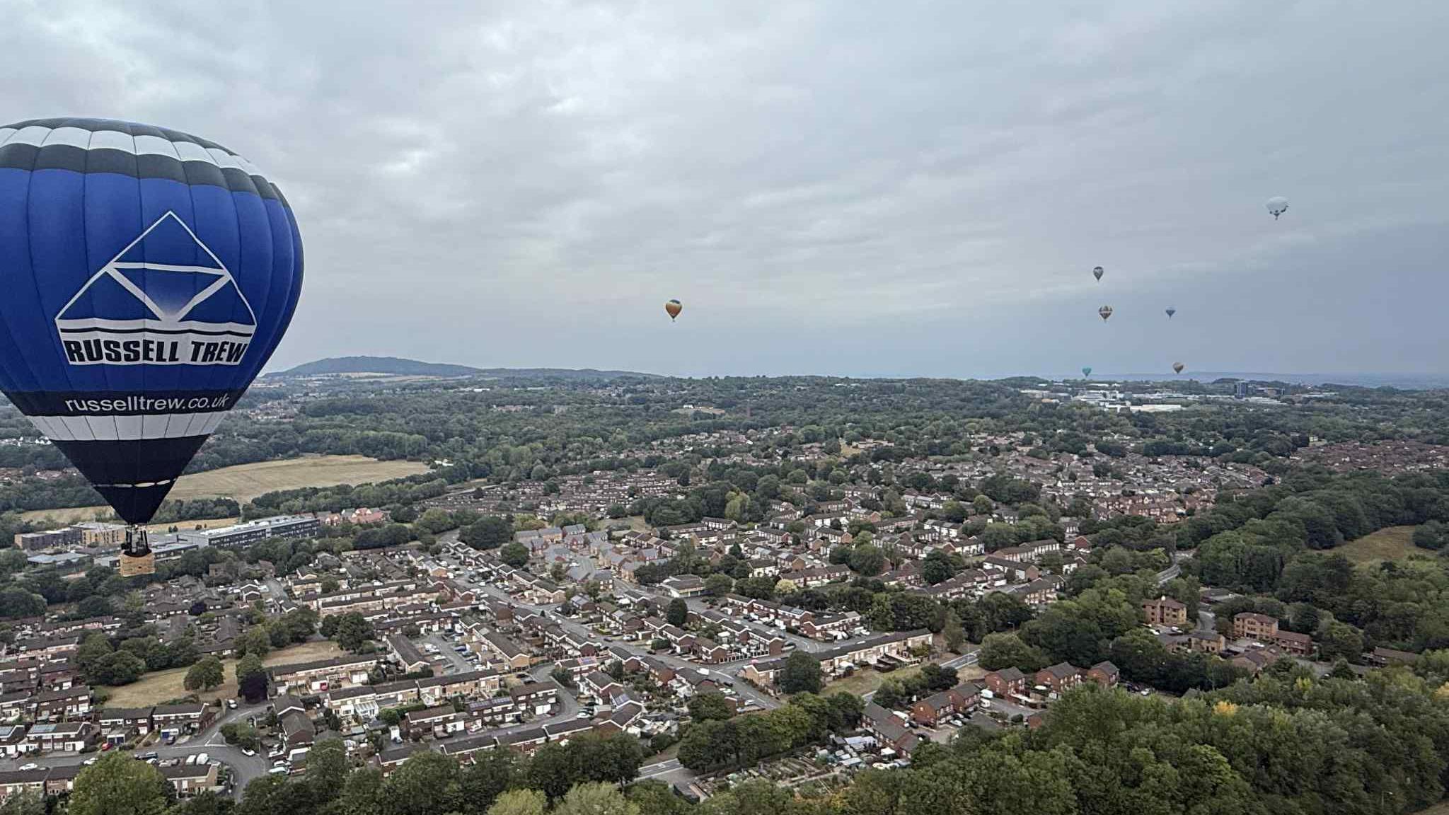 View of multi-coloured hot air balloons in the sky while trees and buildings are below. The skyline looks grey due to the weather and there is a large blue and white hot air balloon that has the words "Russell Trew" in black