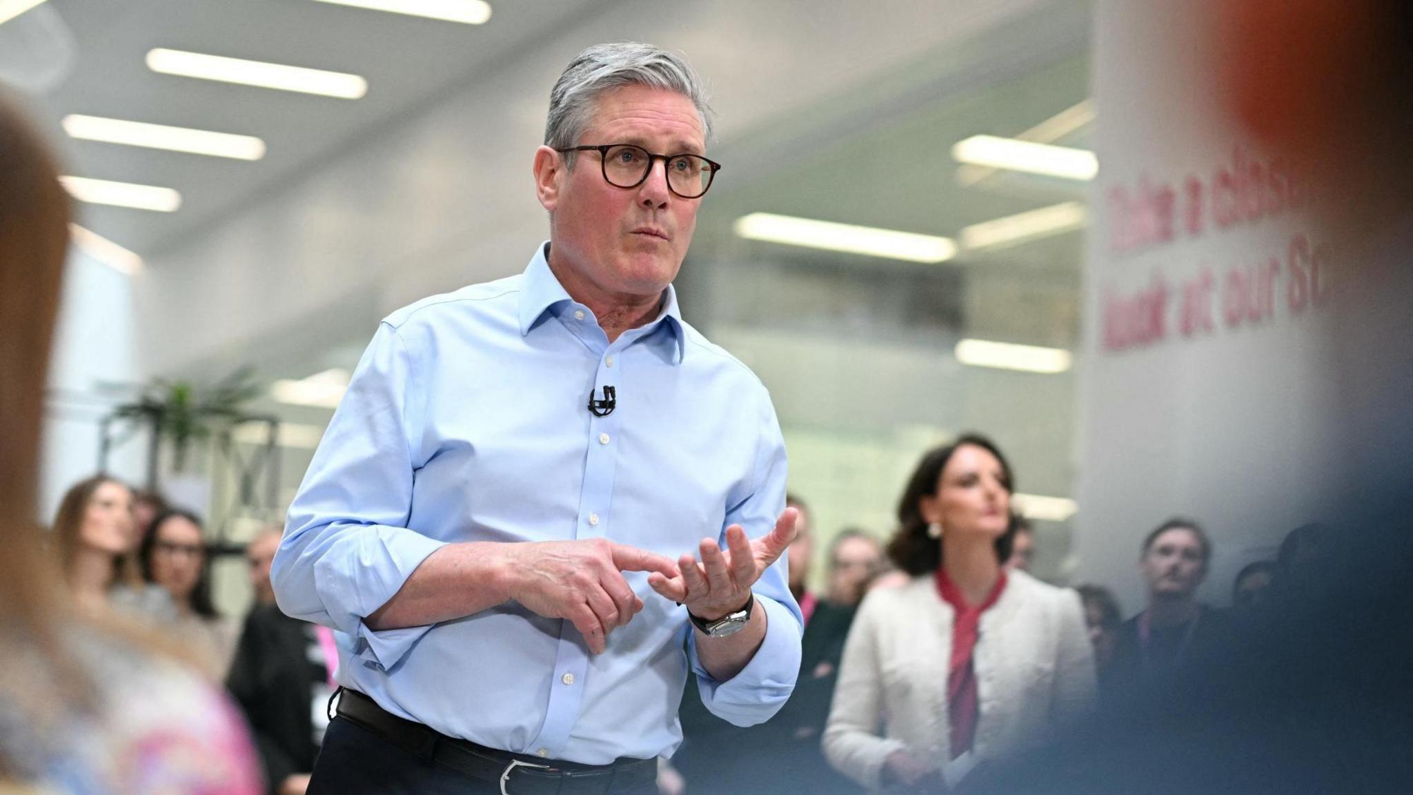 Keir Starmer delivers a speech at a government building. He's wearing a blue shirt and black glasses.