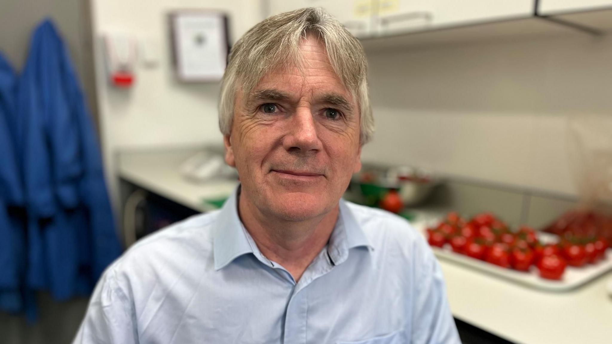 Martin Warren sits on a chair in front of a workbench where tomatoes are being prepped. He is wearing a pale blue shirt and has grey hair. He is looking at the camera and is smiling. You can see his head and shoulders. There are rows of tomatoes on a tray behind his left shoulder. To his right there is a rail with blue lab coats hanging up.