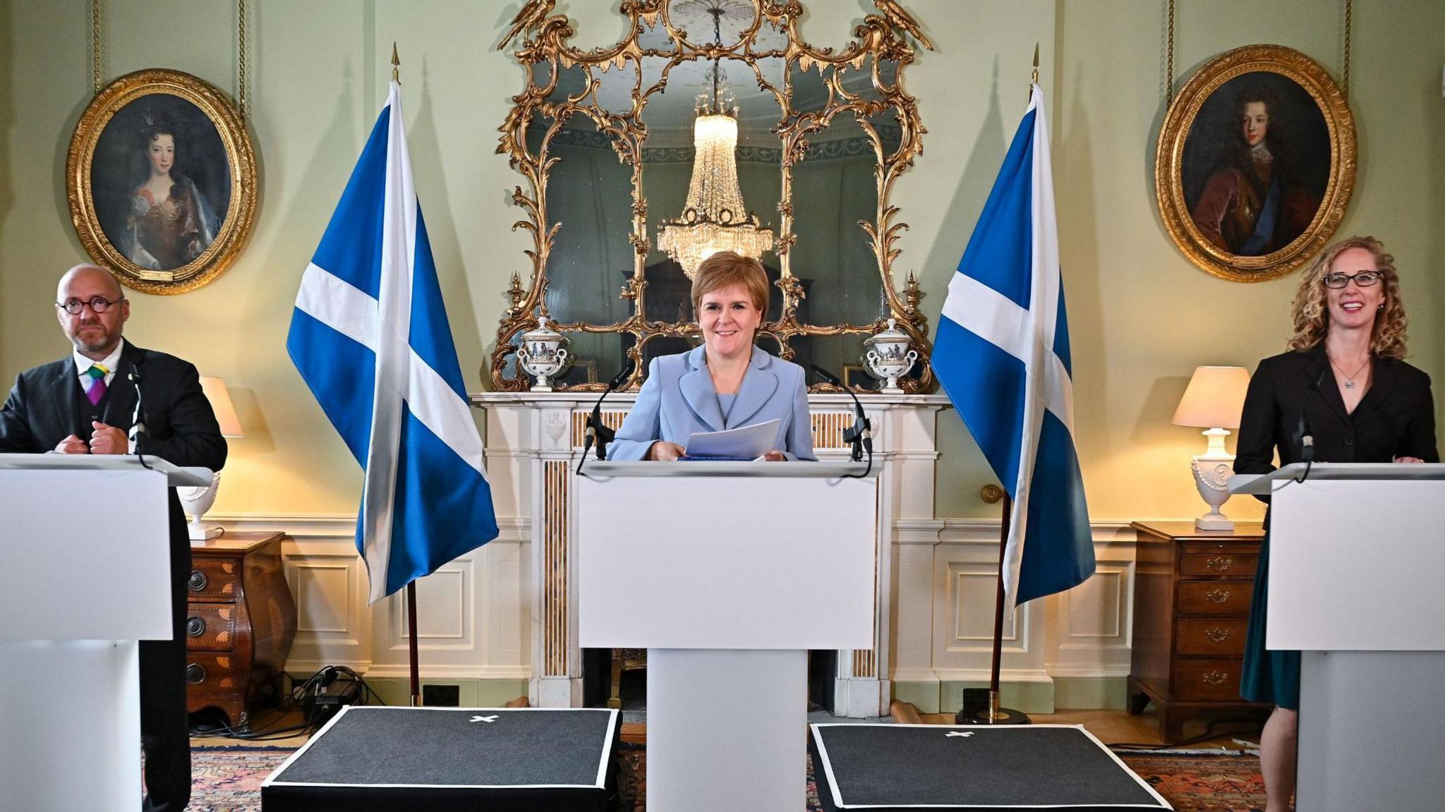 A bald man in a dark suit stood behind a rostrum and to his left is a woman in pale blue suit stood behind a rostrum, and to her left is a woman in a dark dress stood behind a rostrum