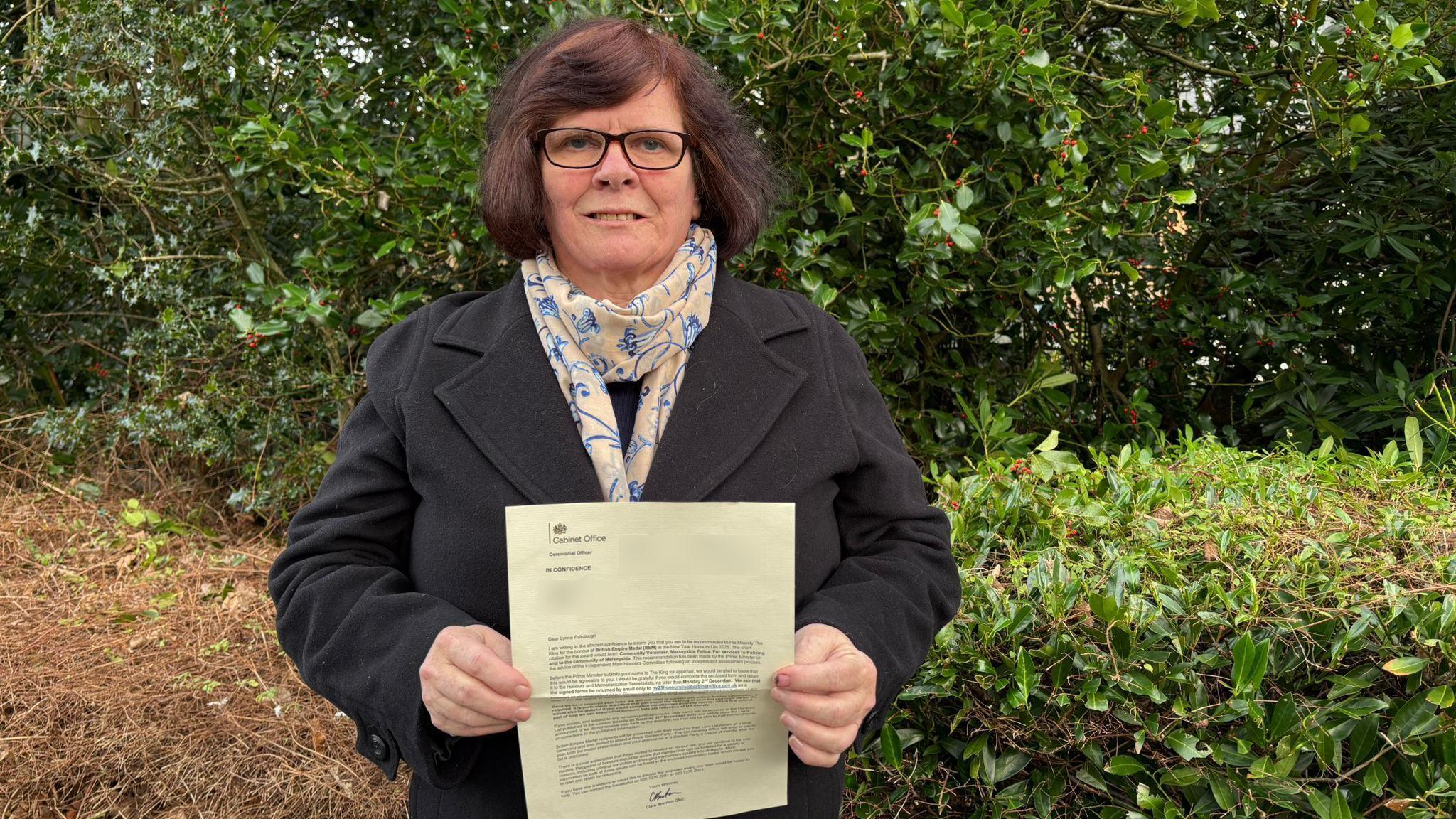 Lynne Fairclough, with brown hair and wearing glasses, smiles at the camera while holding a copy of a letter from the Cabinet Office