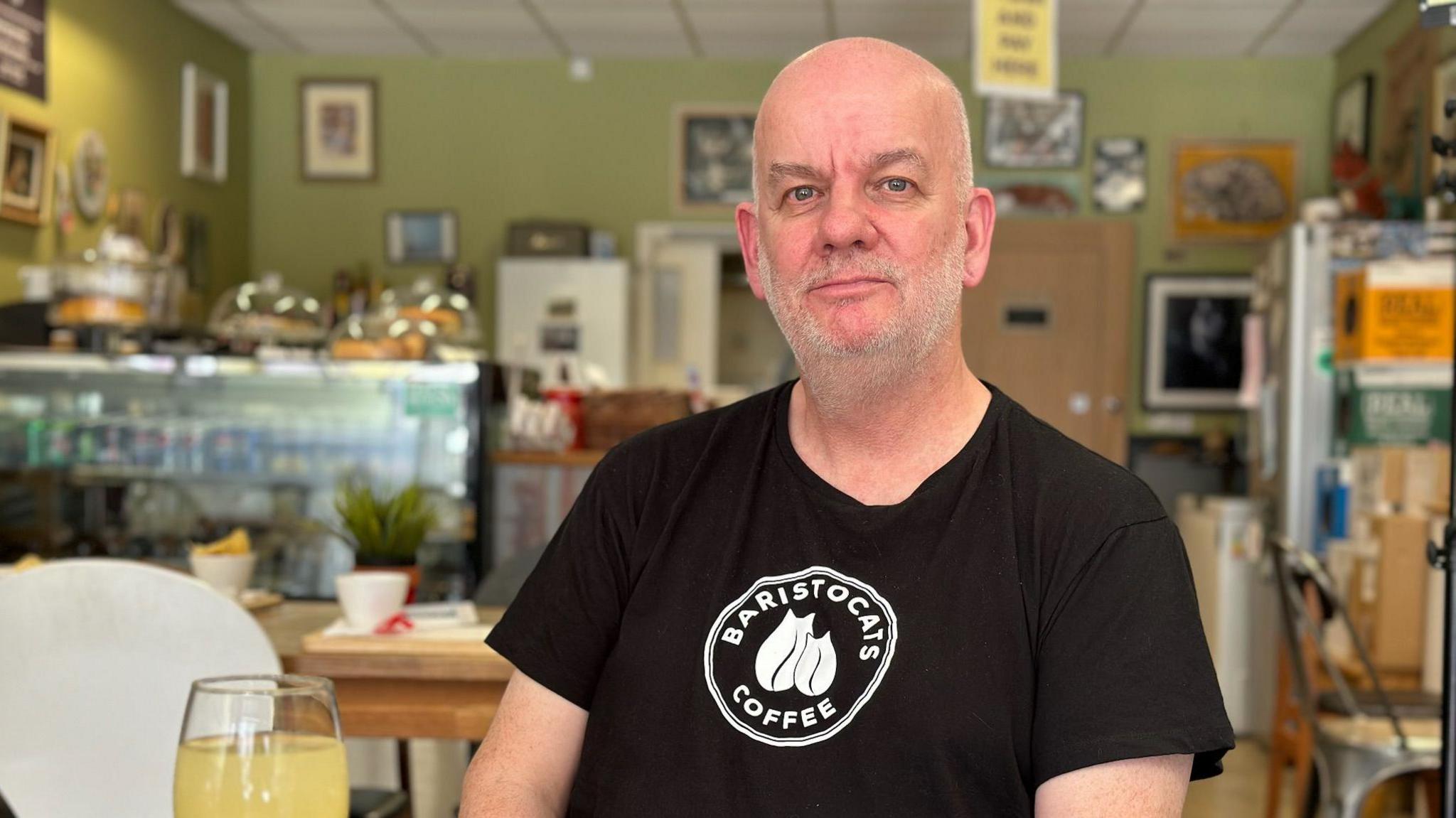 Marcus Kittridge sitting in his cafe wearing a black T-shirt that says Baristocats Cafe on it, with a glass of orange juice on the table in front of him