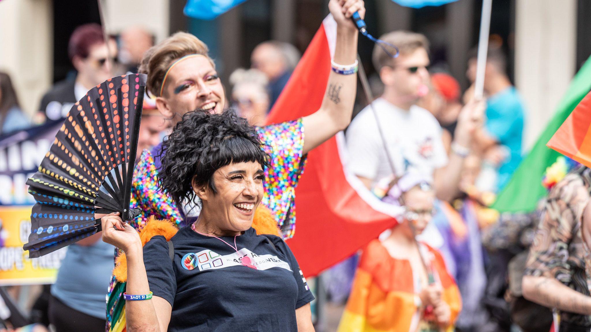 A woman in a black t shirt holds a rainbow fan. She is smiling broadly. Behind her a man in a sequinned top raises and arm in celebration. Other colourful banners and costumes can be seen on people around them, blurred in the background.