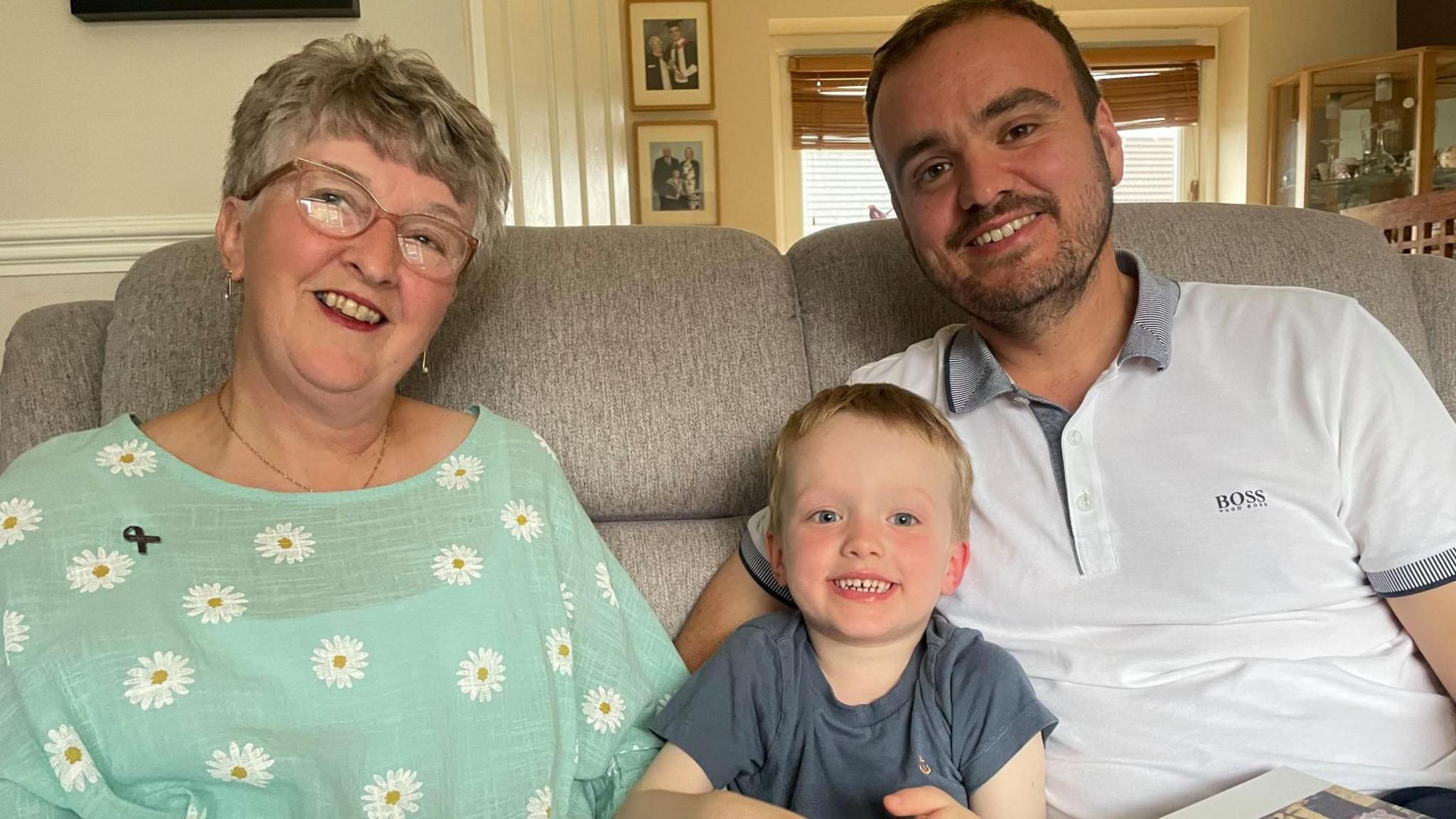 An older woman, a young boy and a middle-aged man pose together on the sofa smiling