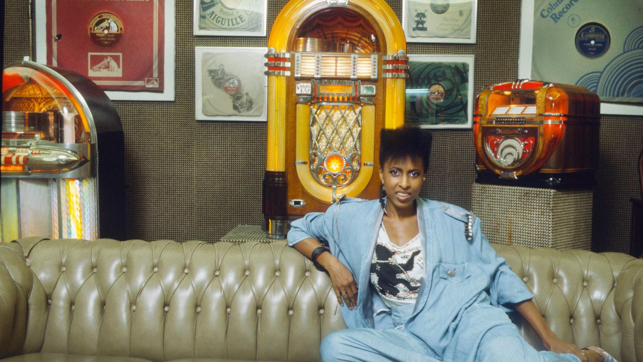 Singer Nona Hendryx - a woman with short black hair wearing a blue suit - posing with a Wurlitzer jukebox in 1985.
